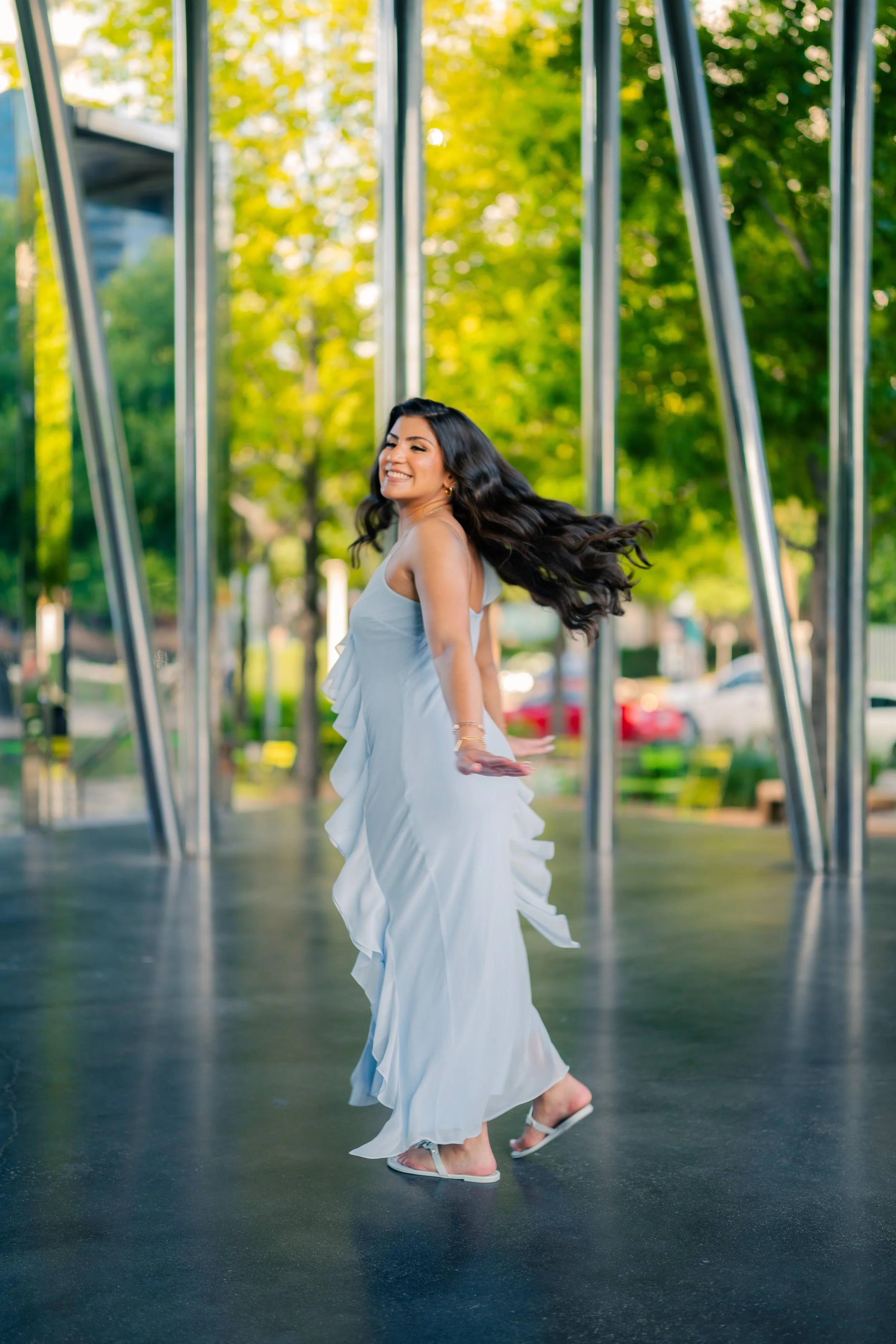A woman in a long white dress with ruffles is dancing outdoors on a black surface, with many trees and some parked cars in the background.