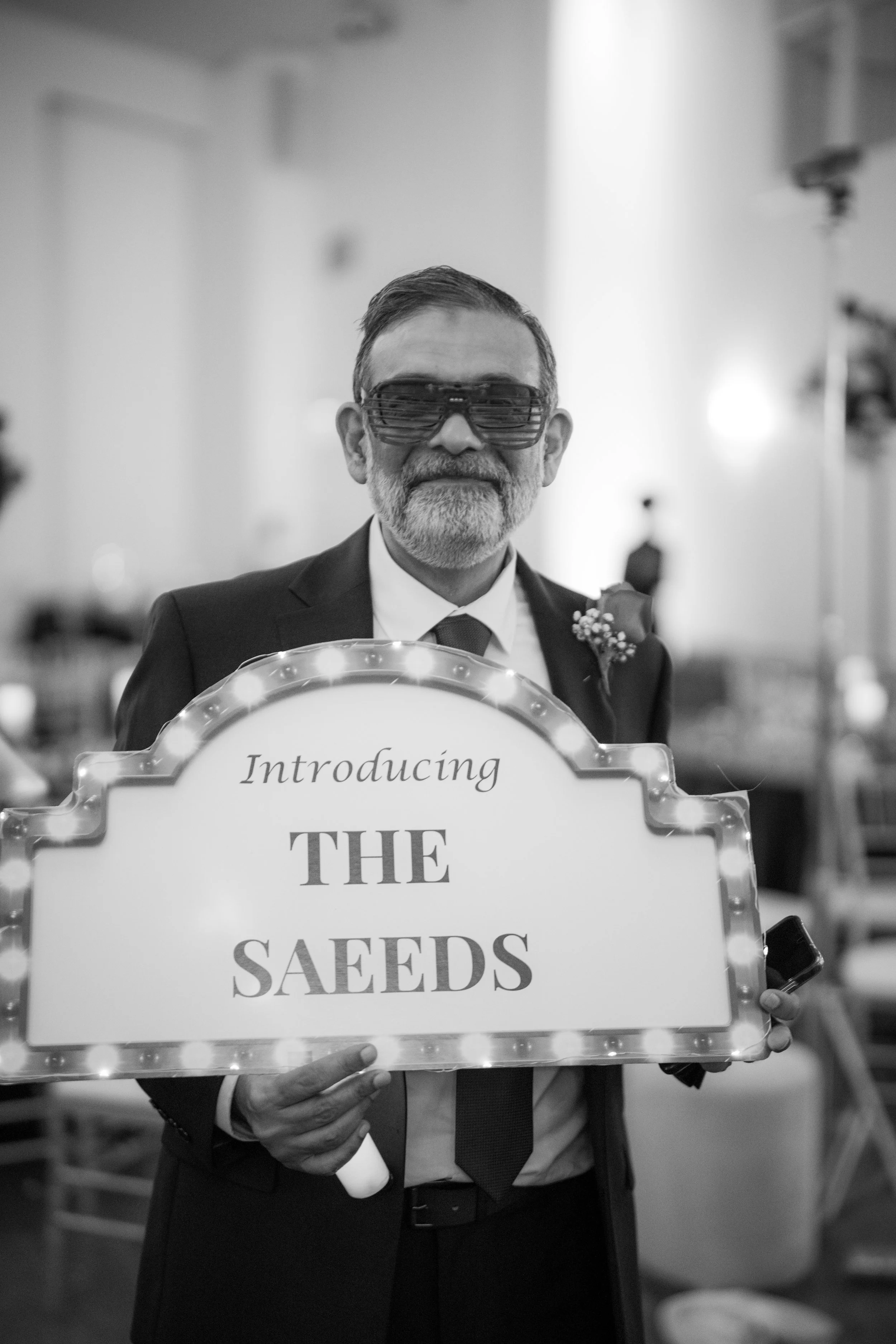 A man wearing a suit and sunglasses, holding a sign that says "Introducing the Seeds," at an indoor event.