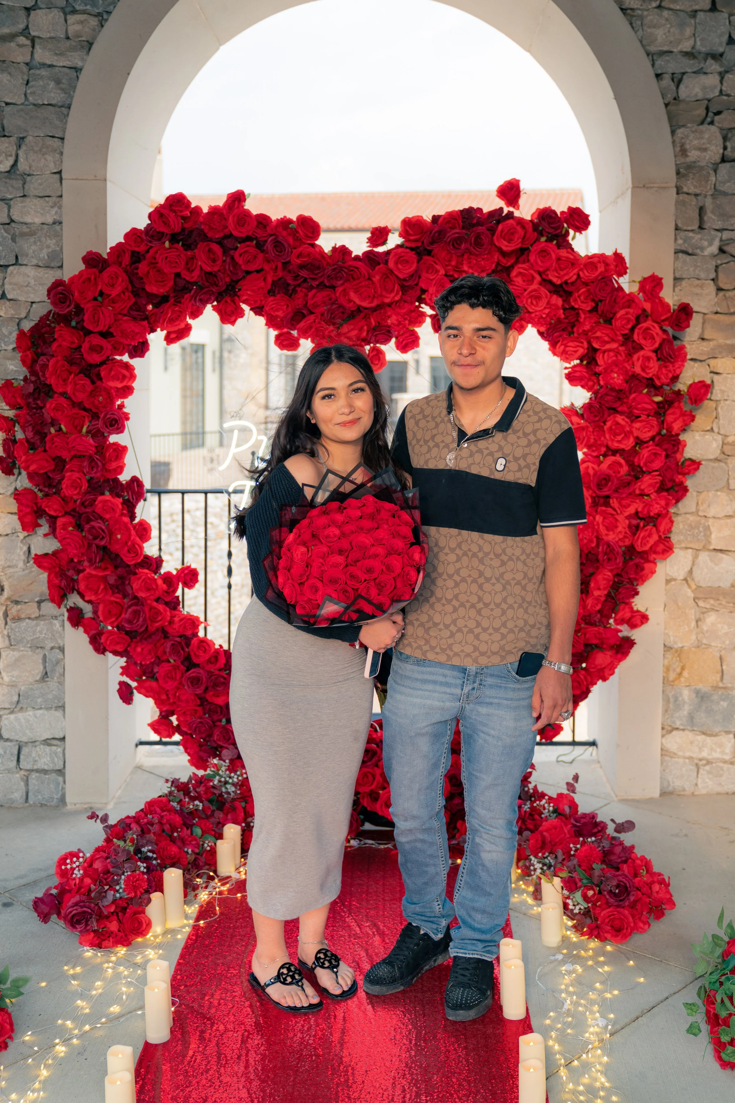 A young couple standing on a red carpet with a heart-shaped rose decoration in the background, holding a bouquet of red roses, celebrating a romantic occasion.