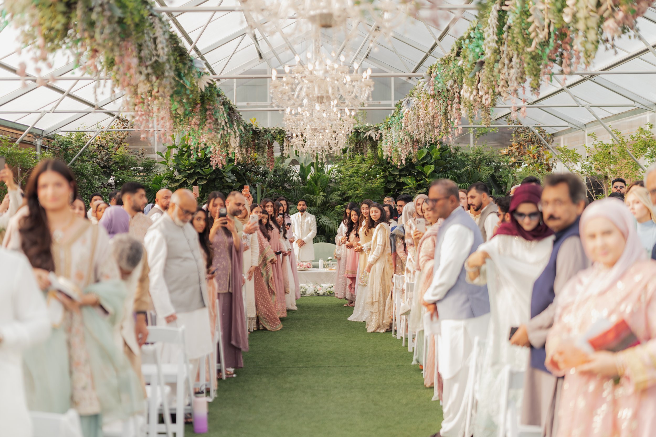 Wedding guests standing in rows at a wedding ceremony inside a glass greenhouse, decorated with hanging flowers and a sparkling chandelier.