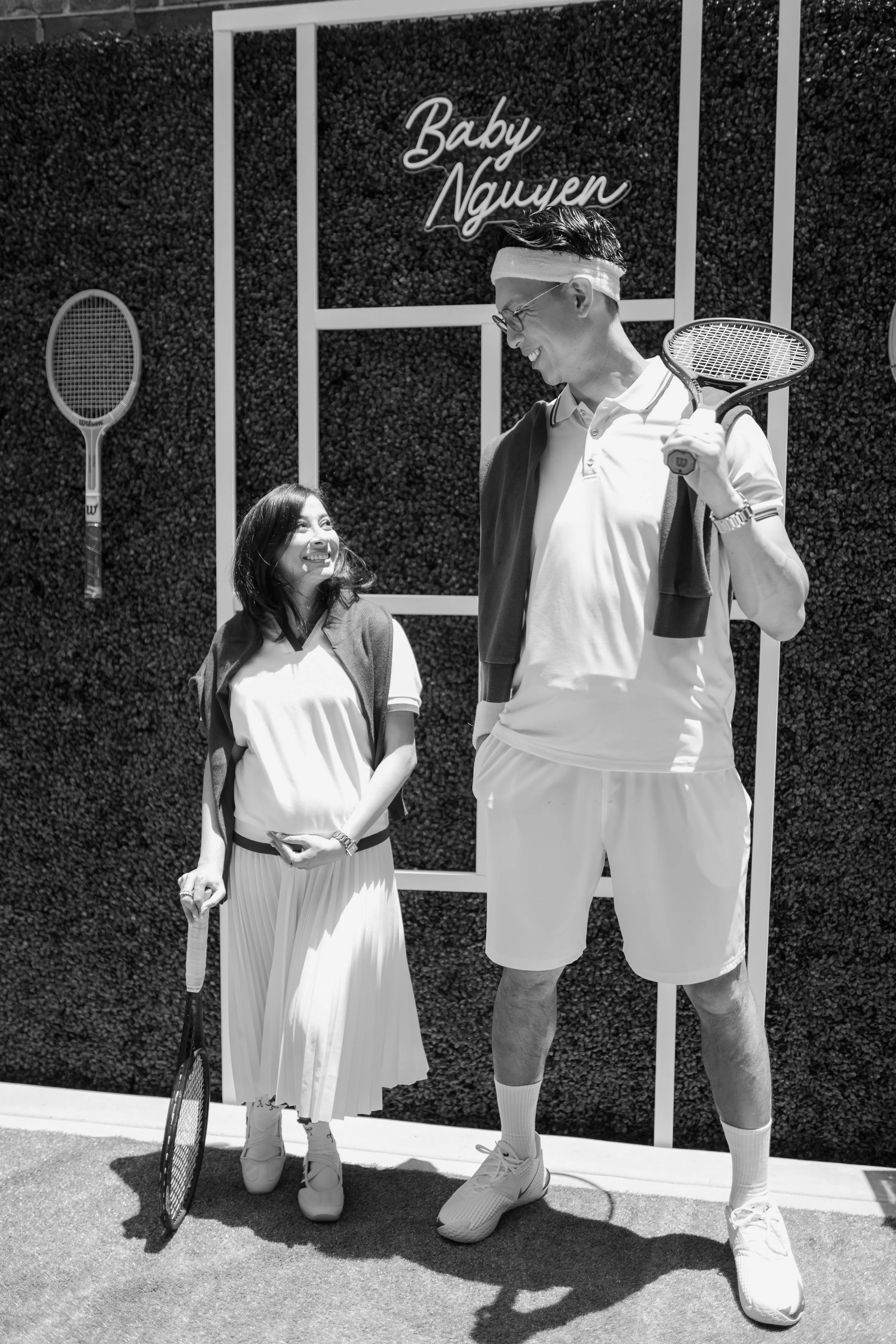 A man and woman are standing outdoors near a hedge. The man is holding a tennis racket over his shoulder and wearing a headband, glasses, a polo shirt, shorts, and sneakers. The woman is holding a tennis racket, smiling, and wearing a pleated skirt a
