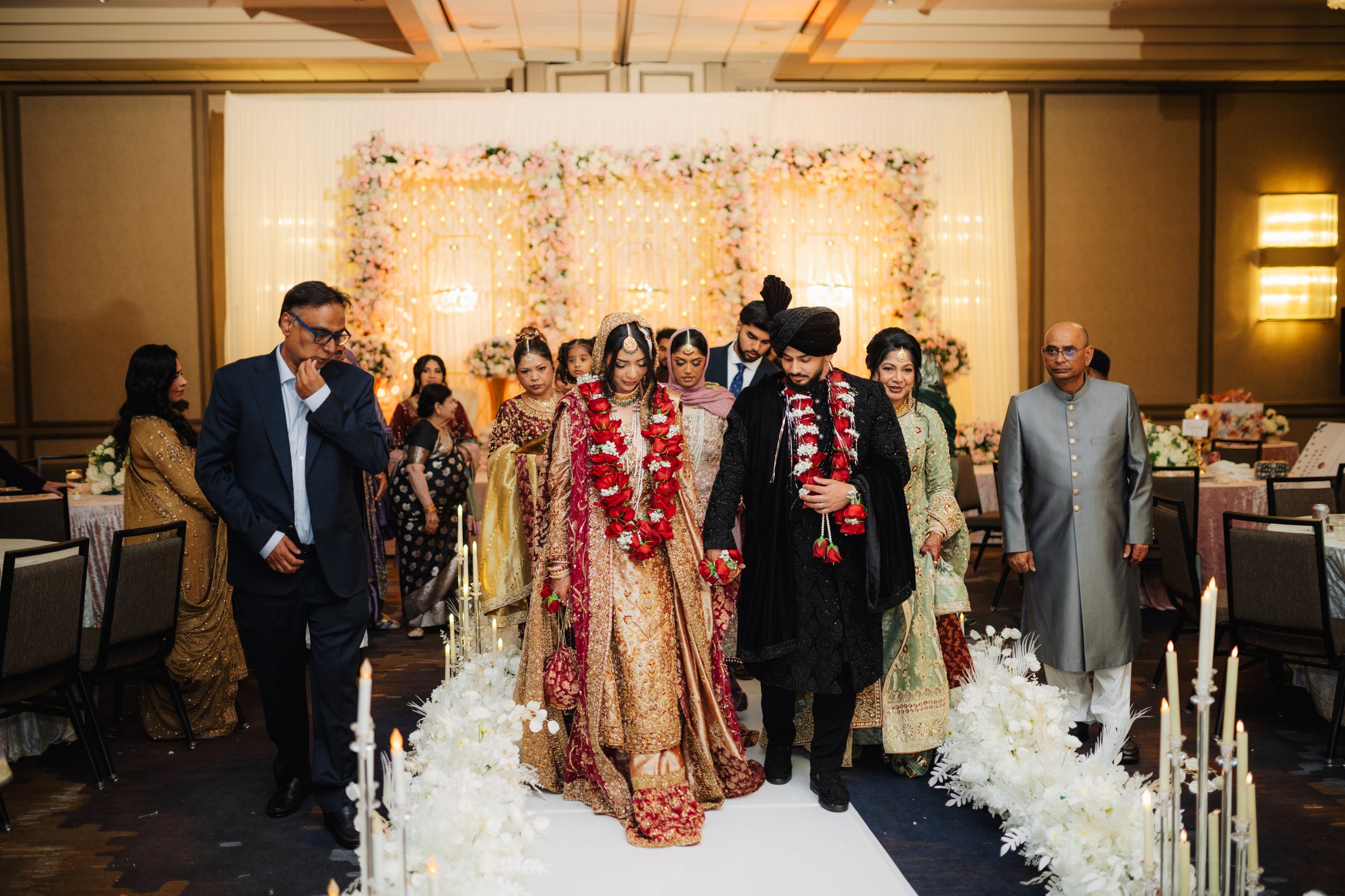 A group of people dressed in traditional and formal attire at a wedding ceremony in an indoor venue decorated with pink and white flowers and fairy lights.