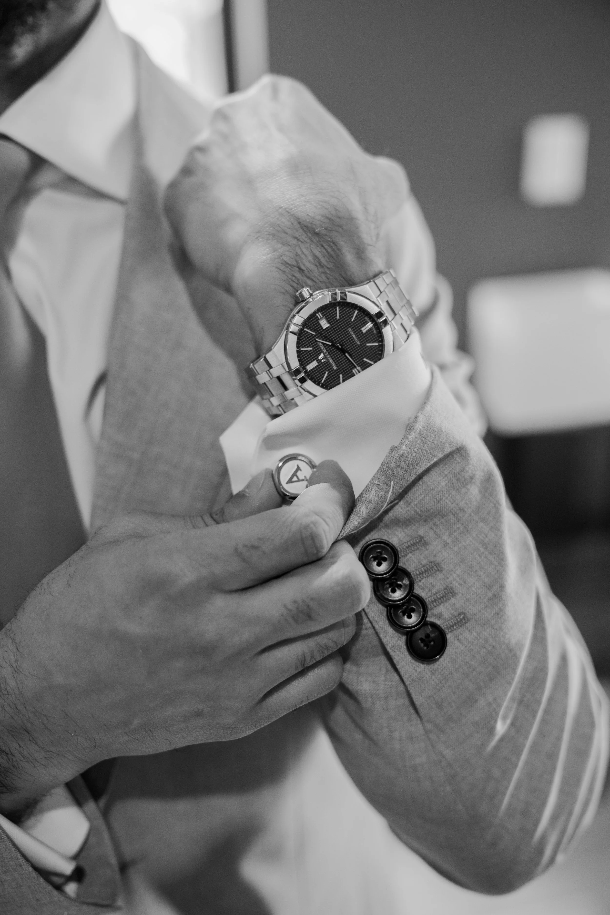 A person wearing a light-colored suit with dark buttons on the sleeve, adjusting a wristwatch on their left wrist. The watch has a metal band and a dark face. The person's hand is holding a button on the sleeve of their jacket.