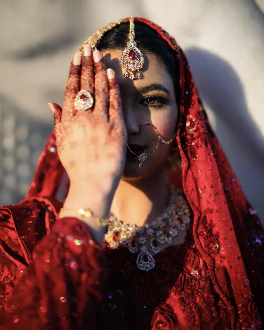 A woman dressed in traditional Indian bridal attire, wearing red clothing and ornate jewelry, partially covering her face with her hand adorned with rings and henna designs.