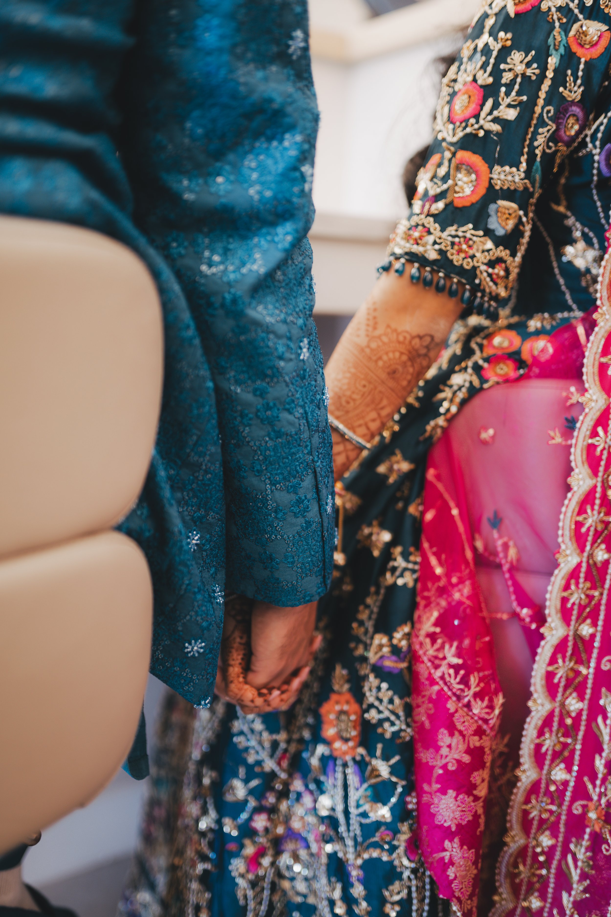 Close-up of women wearing traditional richly embroidered Indian sarees, with visible detailed embroidery and henna on their hands, at an event or celebration.