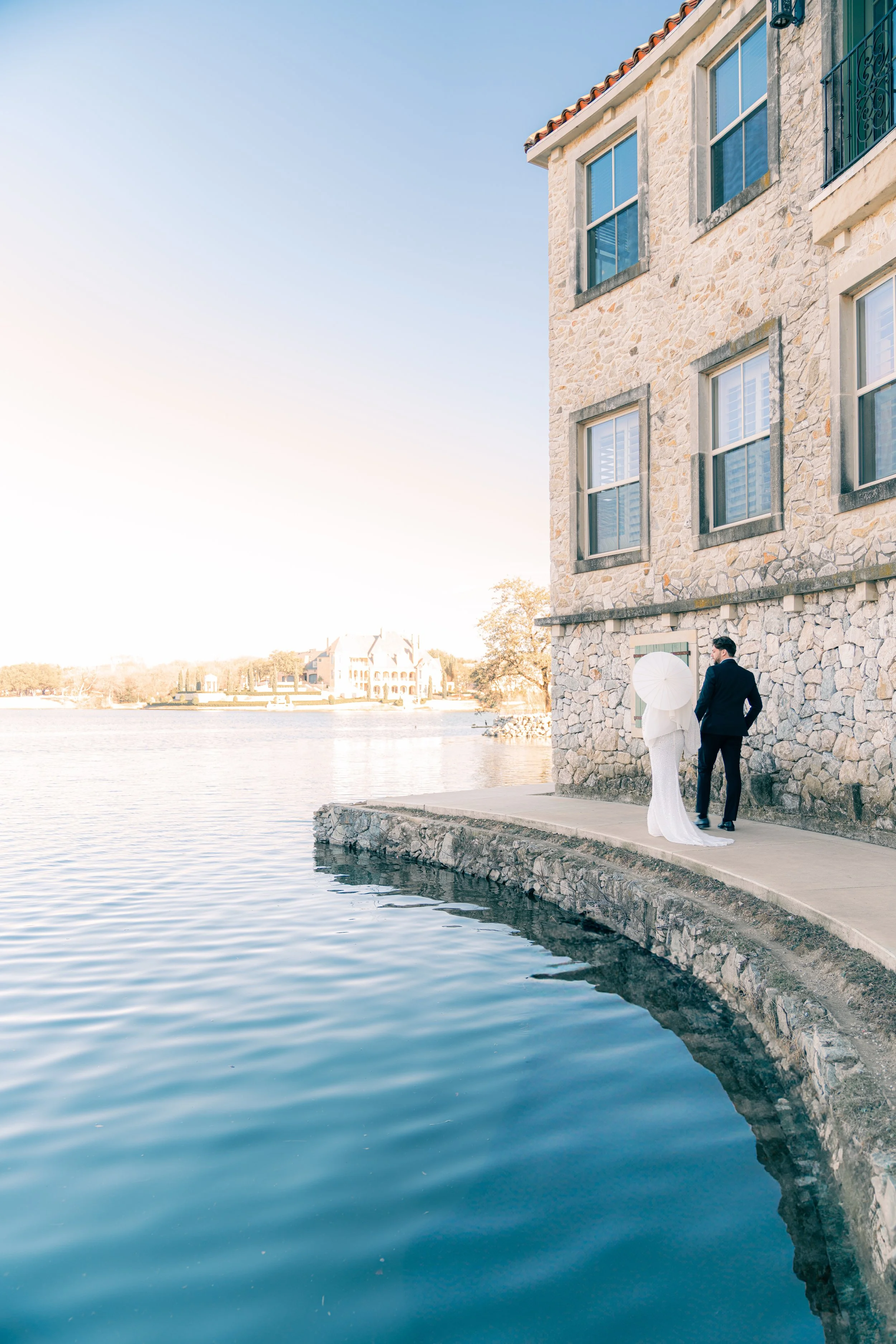 A couple dressed in formal wedding attire standing by a stone building overlooking a body of water, with clear skies and distant houses on the opposite shoreline.
