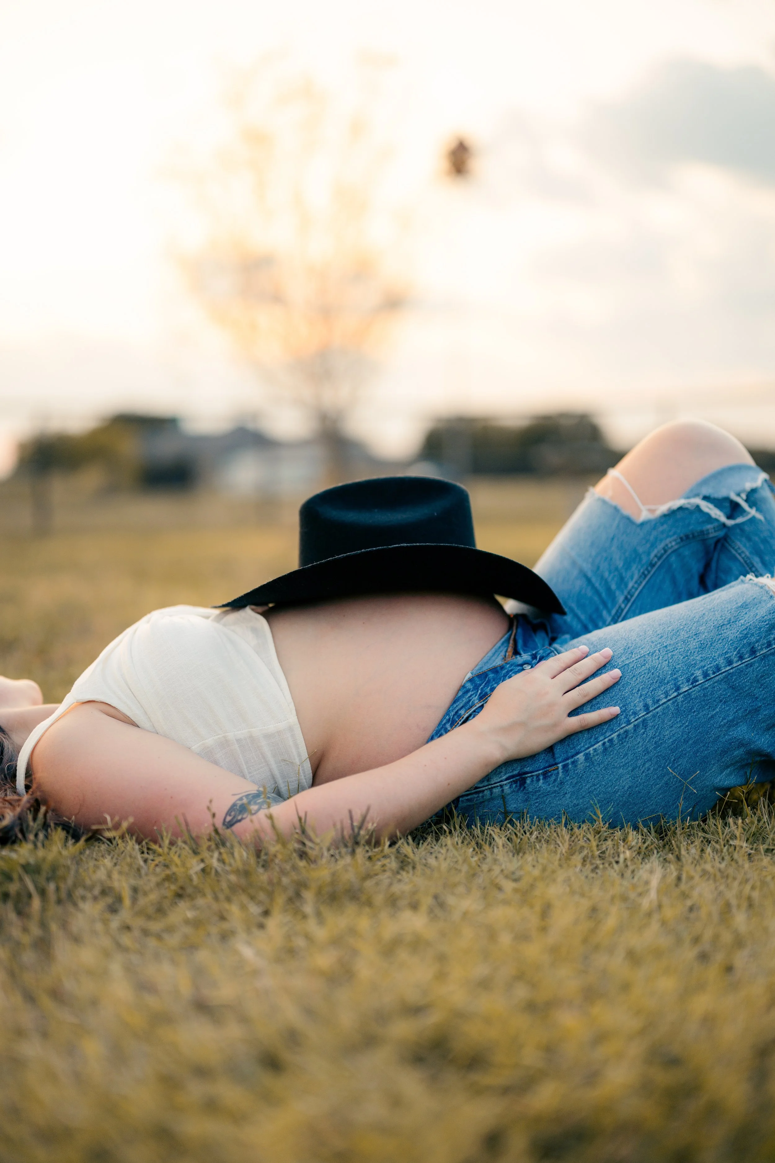 A pregnant woman lying on grass in a field during sunset, wearing a white shirt, ripped jeans, and a black hat. She has a tattoo on her arm and is resting with her hand on her belly.