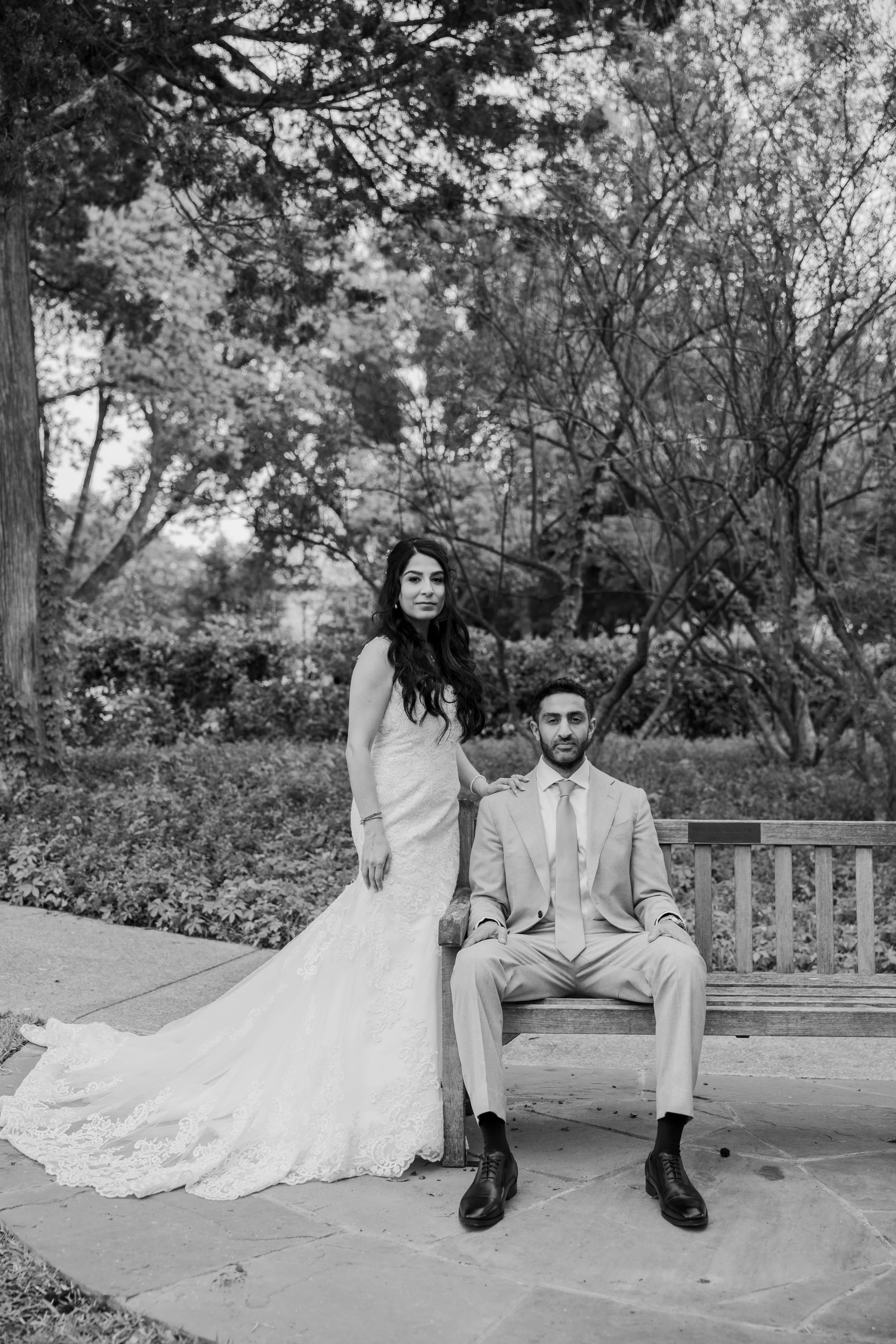Black and white photo of a bride in a wedding gown standing next to a seated groom in a suit, outdoors with trees and shrubs in the background.