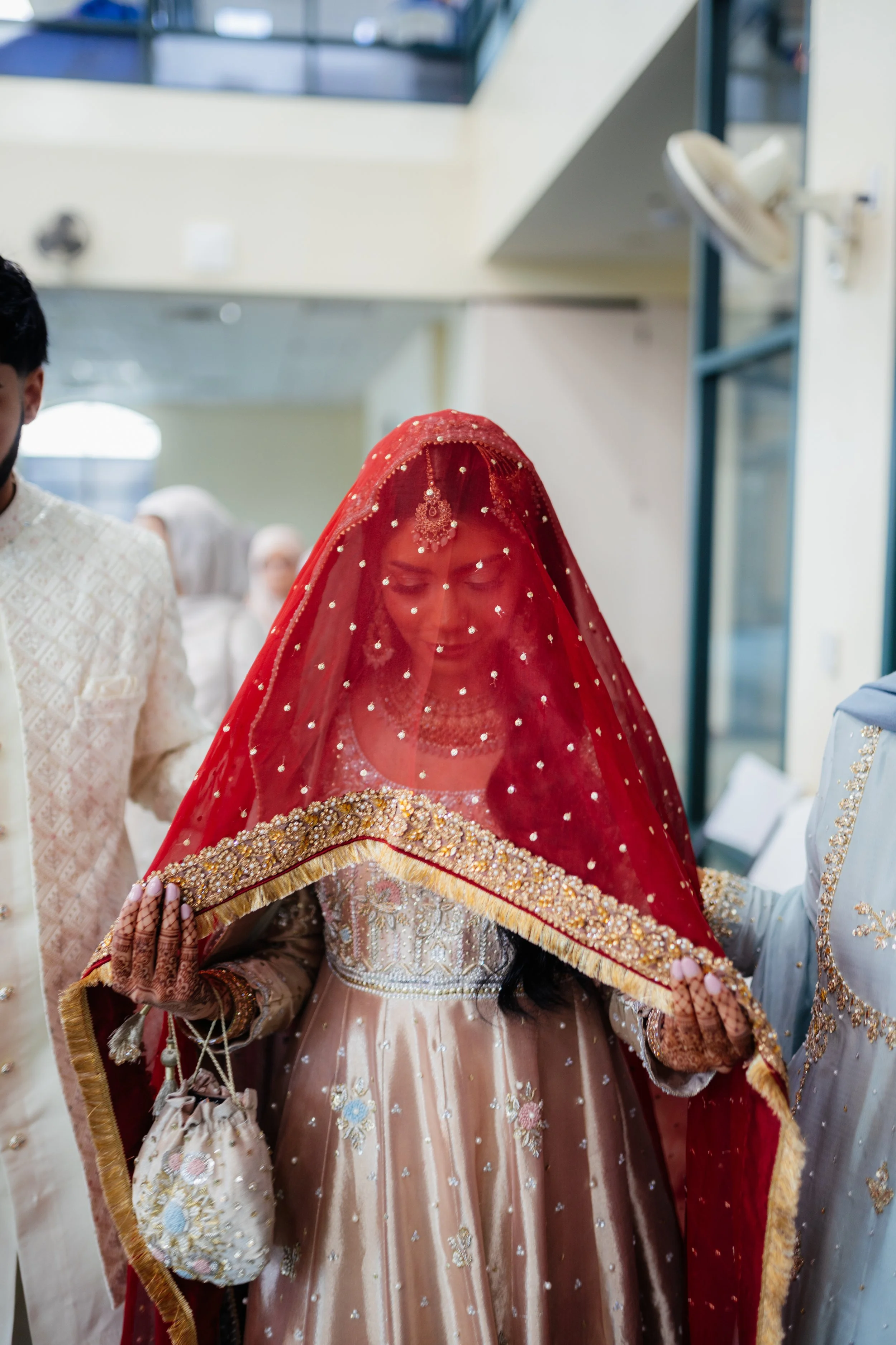 A bride wearing a traditional wedding dress with a veil covering her face, decorated with gold and embroidery, as she prepares for a wedding ceremony.