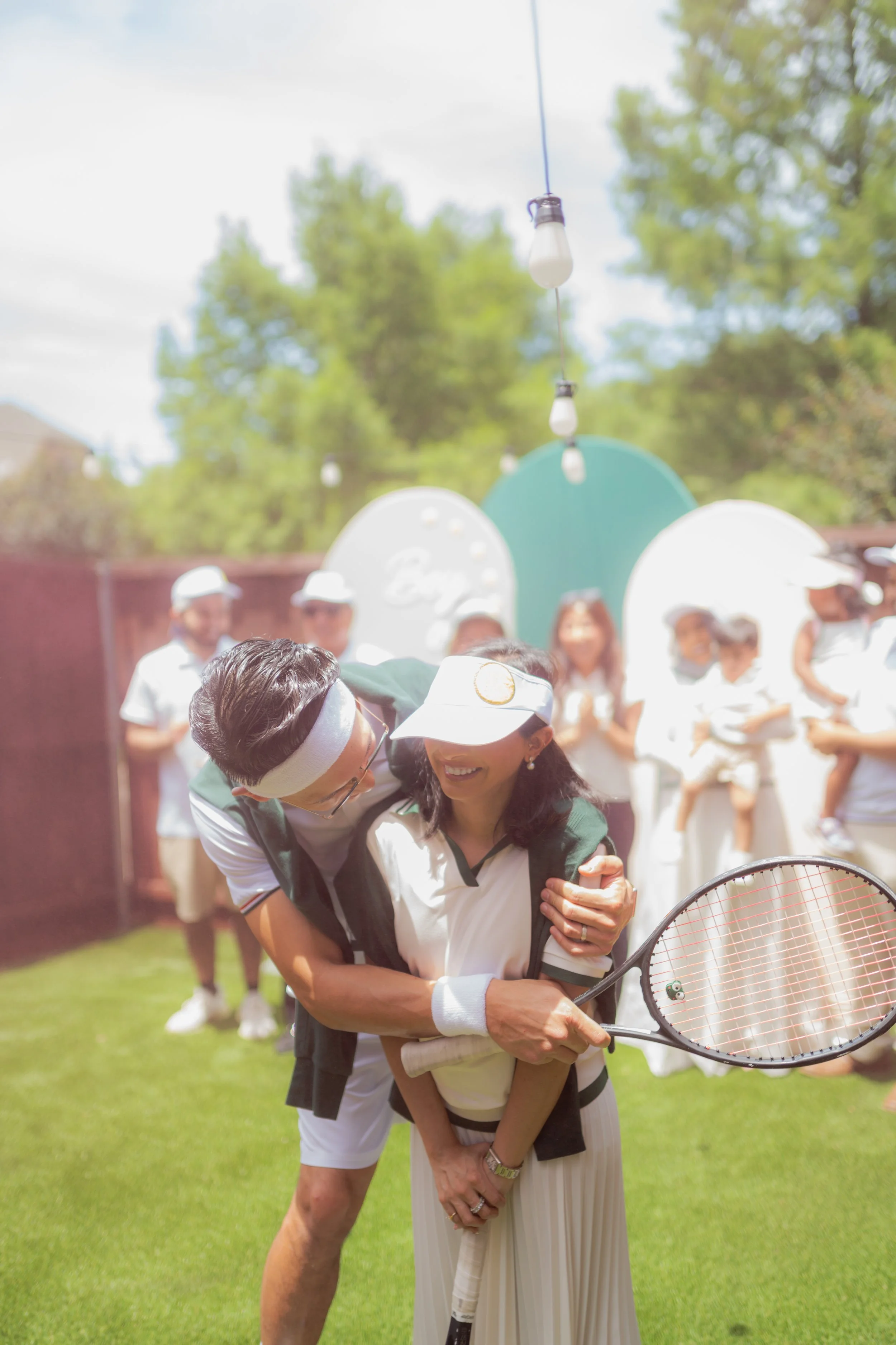 People hugging on a tennis court during a celebration, with a woman holding a tennis racket and a man wearing a headband, both dressed in sportswear.