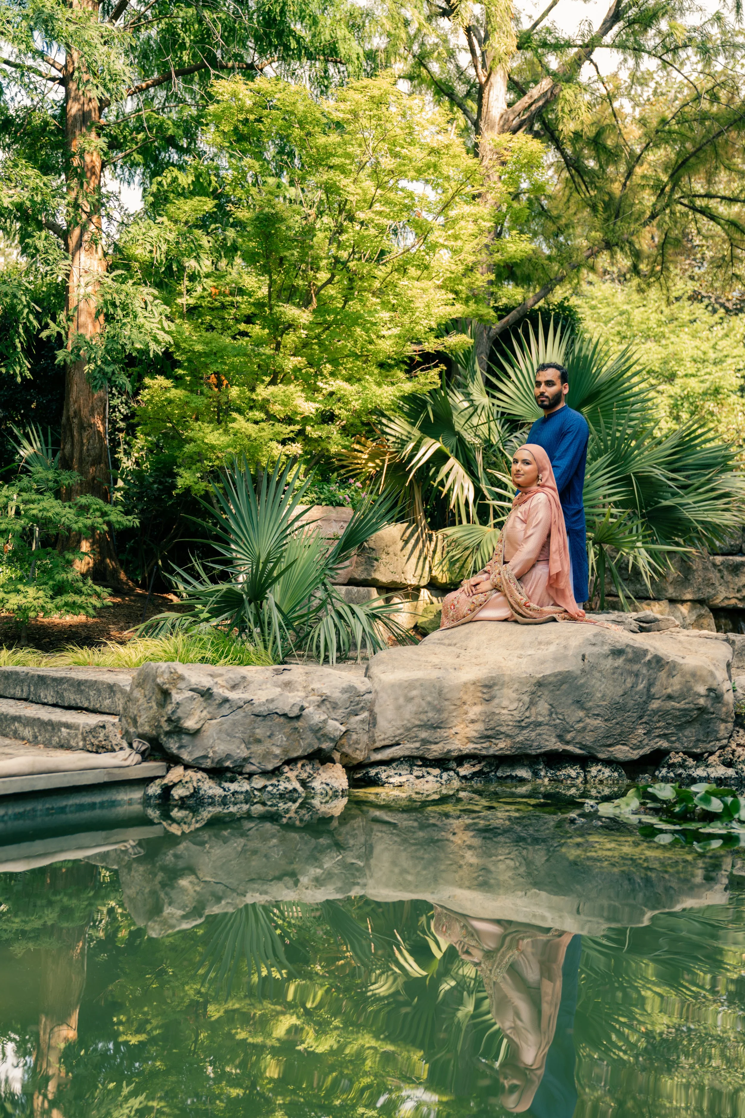 A man and woman in traditional dress are sitting and standing on rocks beside a pond in a lush garden with green trees and tropical plants.