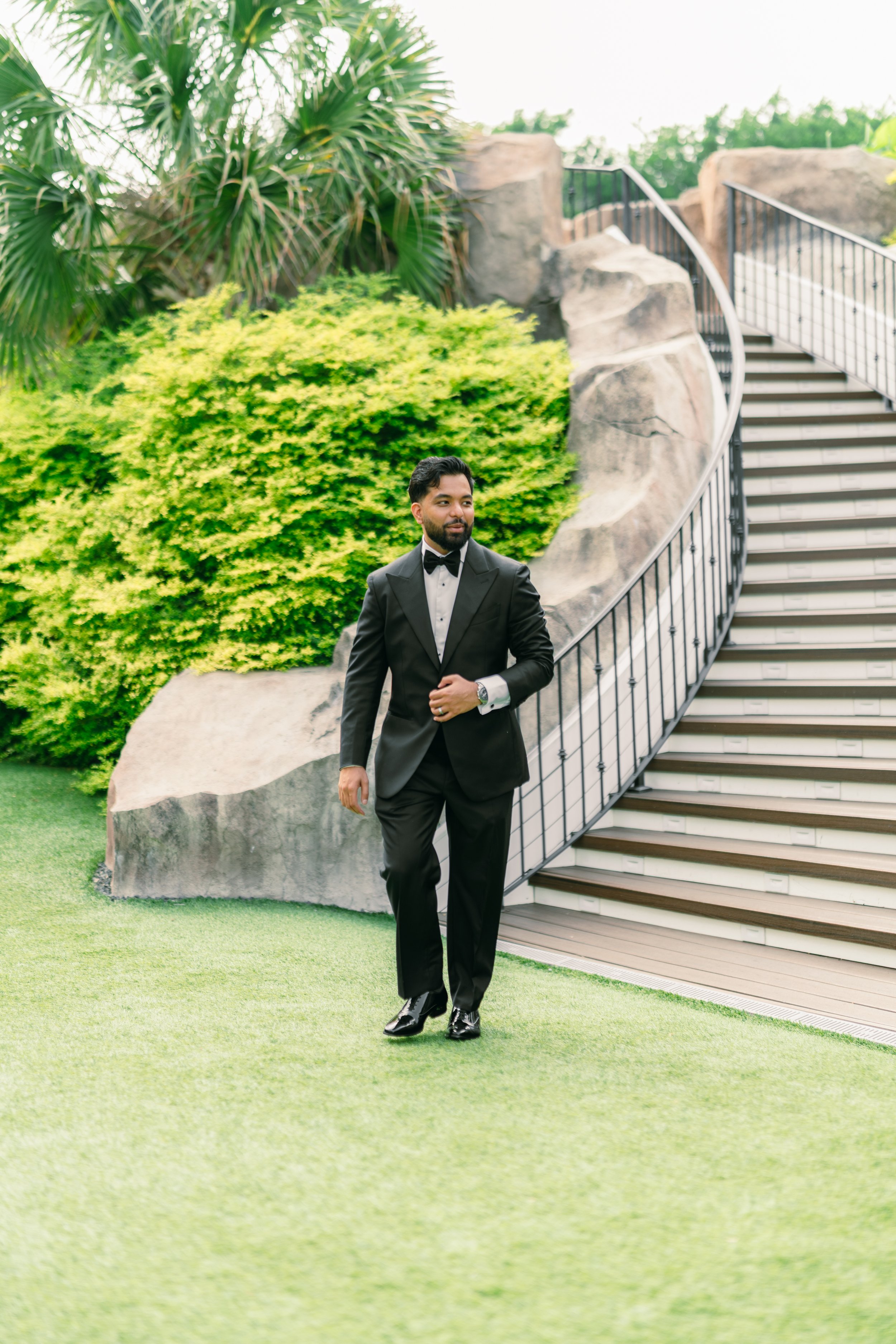 A man in a black tuxedo and bow tie walking on a grassy area near a staircase and lush green bushes.