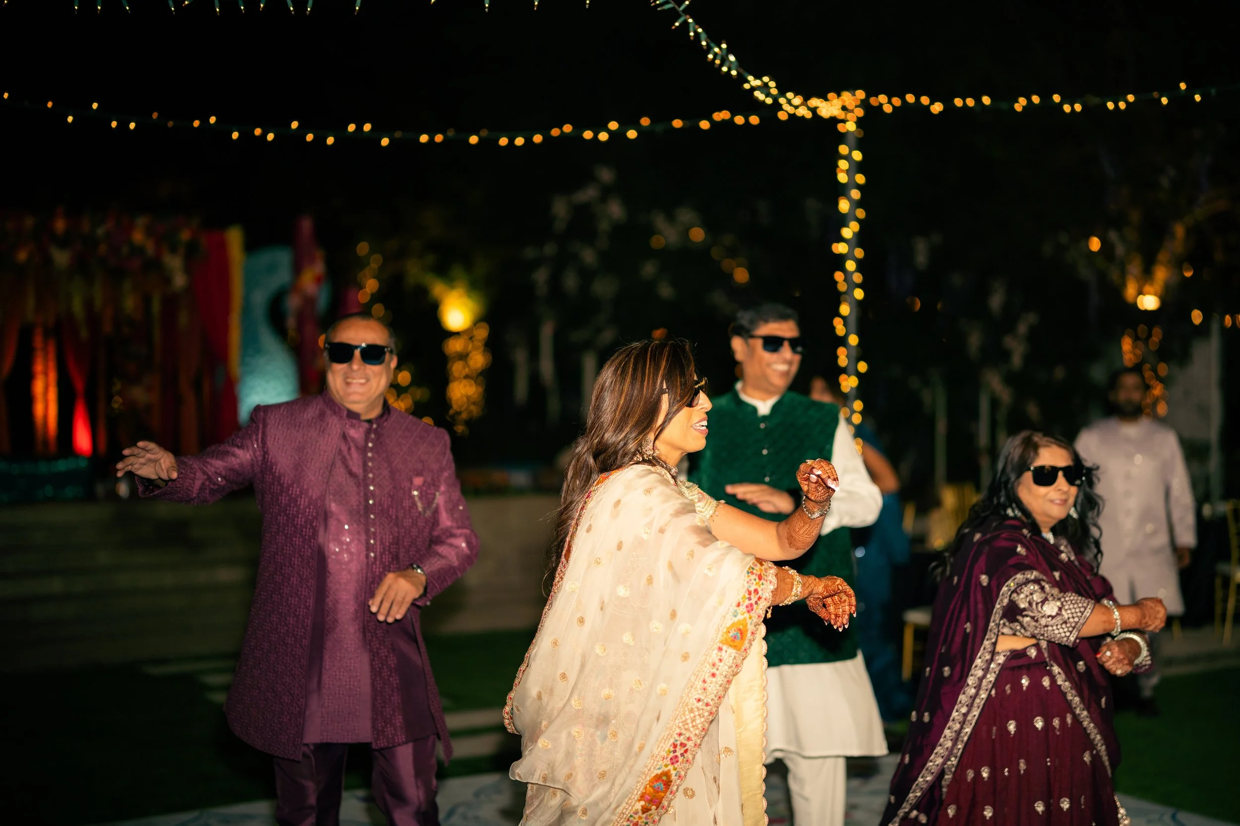 Group of people dancing at an outdoor celebration under string lights, wearing traditional Indian attire and sunglasses.