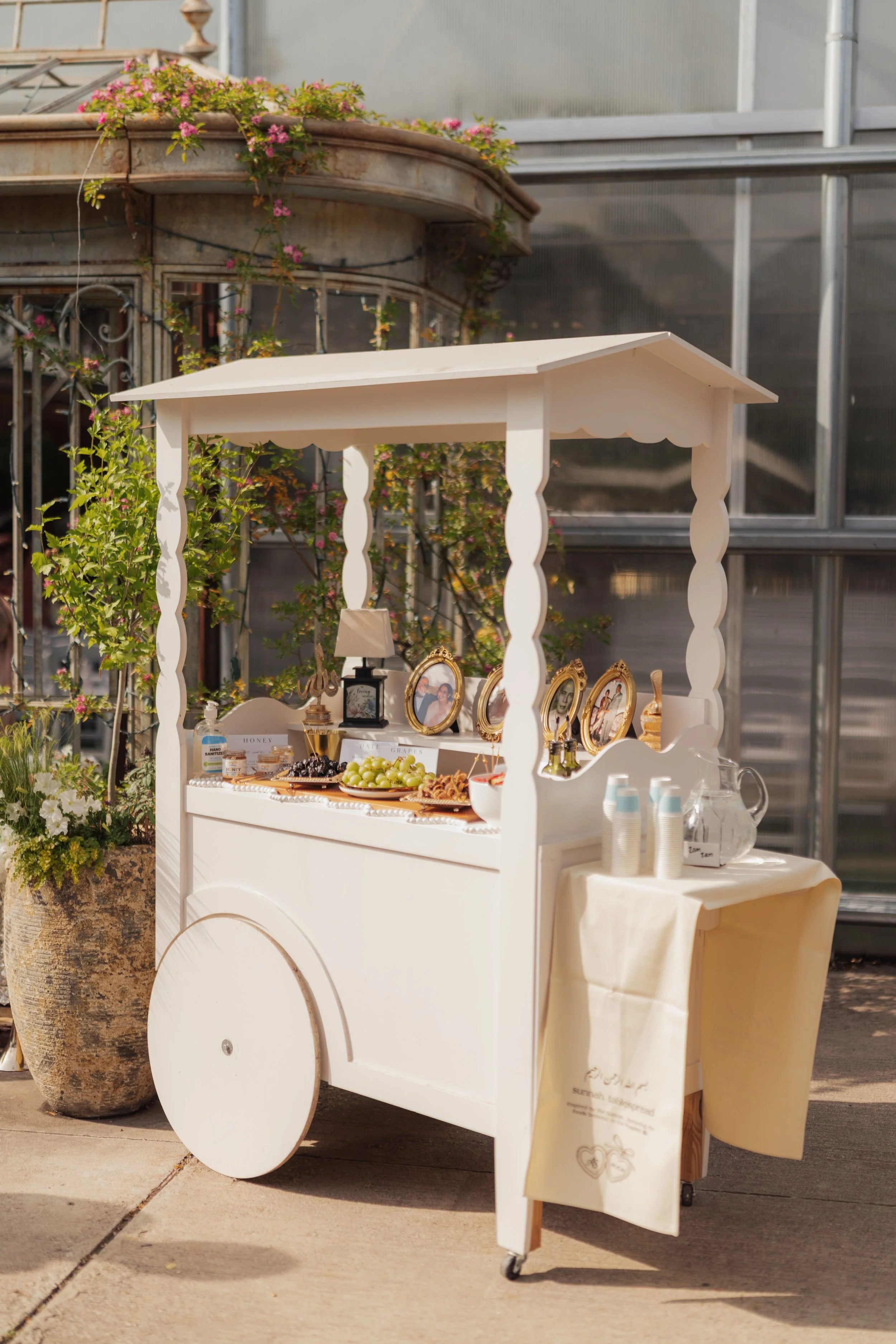 A white dessert or drink cart with decorative elements, flowers, and framed photos on top, situated outdoors with a greenhouse and potted plants nearby.