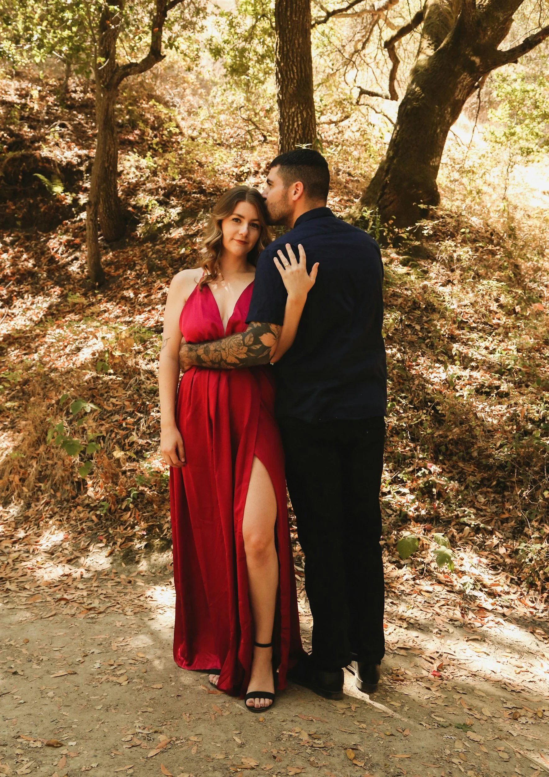 A couple standing on a dirt path in a wooded area, with the man kissing the woman's forehead. The woman is wearing a red dress with a high slit, and the man is wearing a dark shirt and pants. The trees surrounding them are full of green leaves and the sunlight filters through the branches.