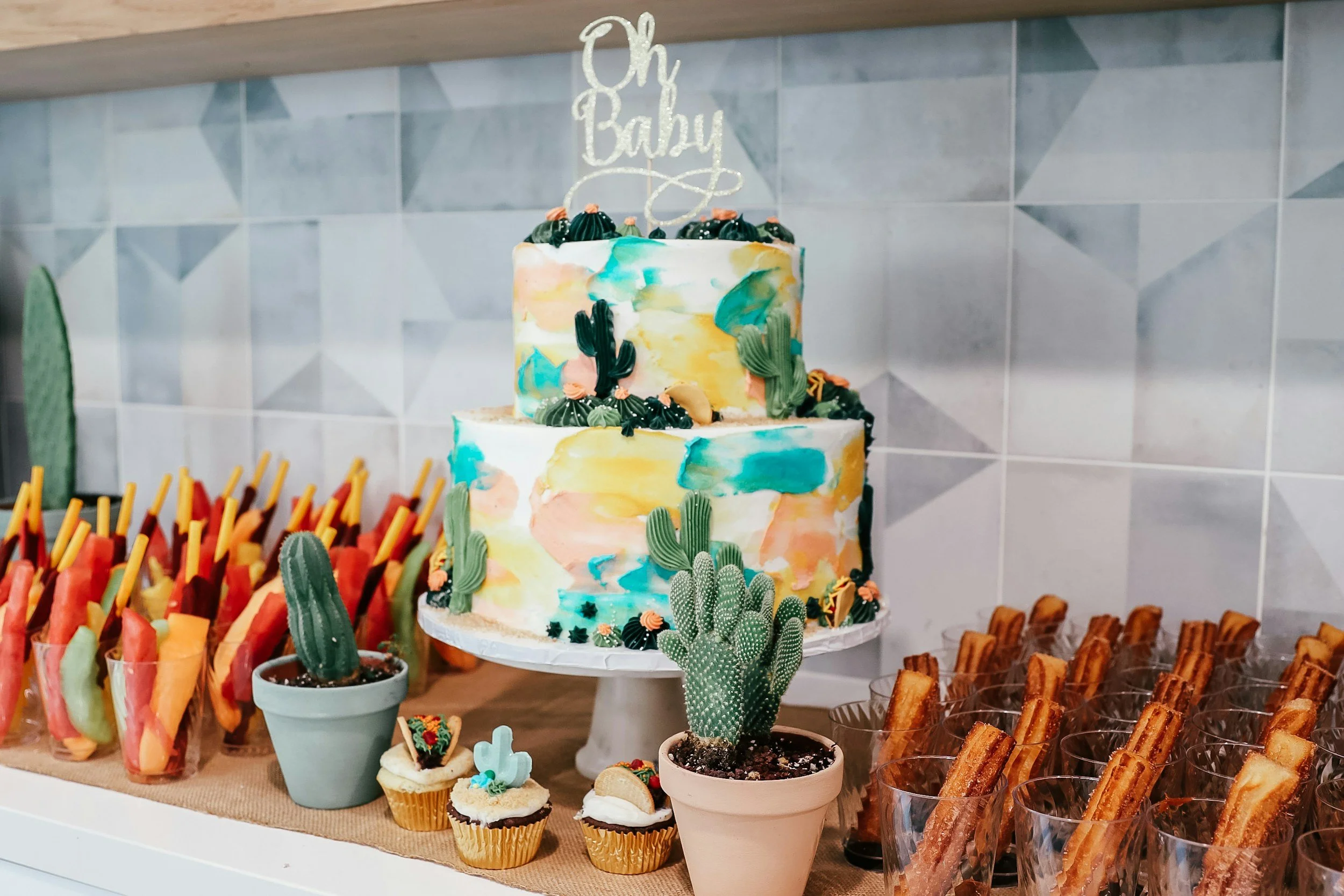 Colorful two-tier cake with cactus decorations, surrounded by churros, cupcakes, fruit skewers, and potted cacti on a table.