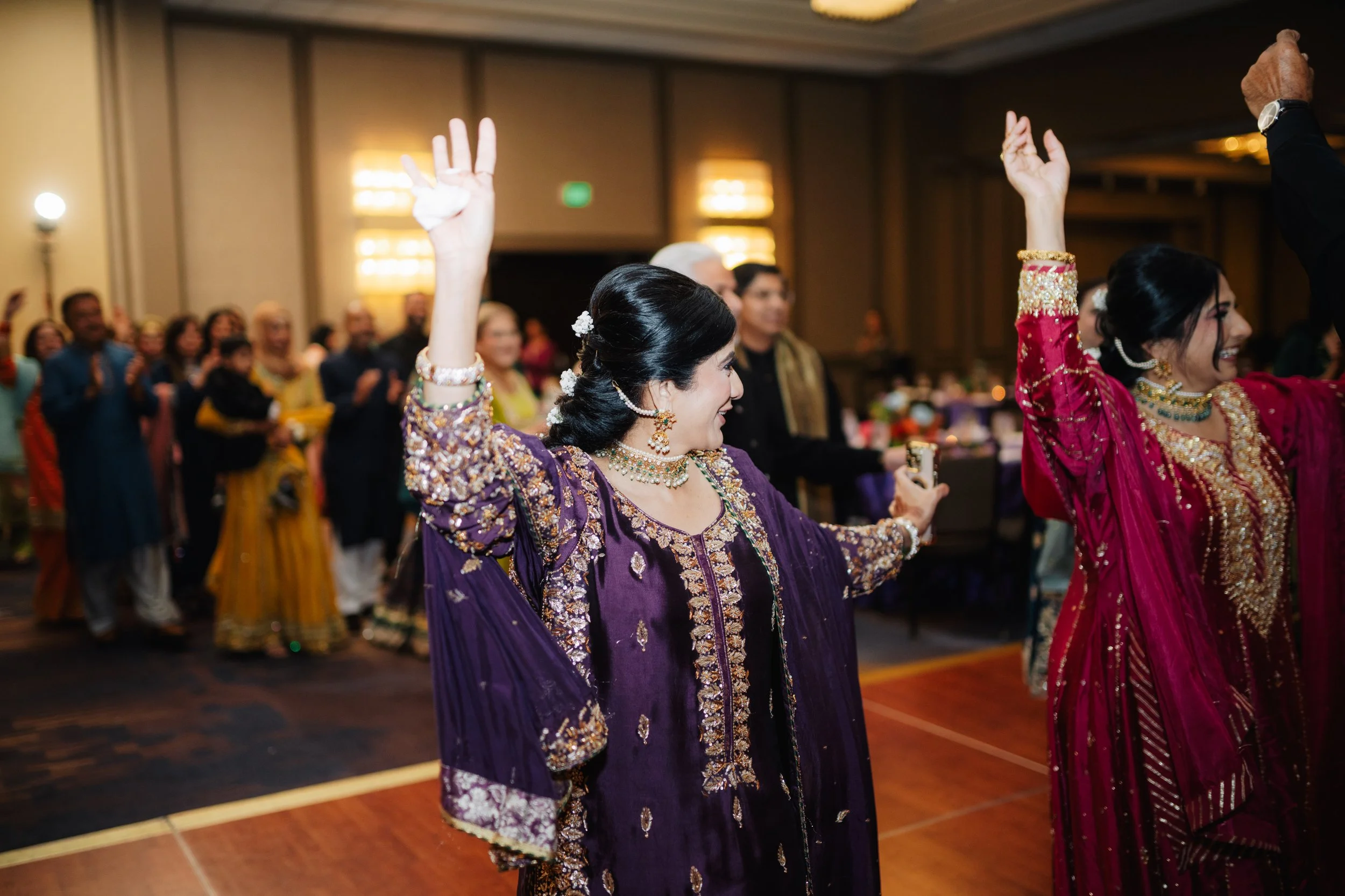 Women in traditional Indian attire dancing at a celebration, with a crowd watching in the background.