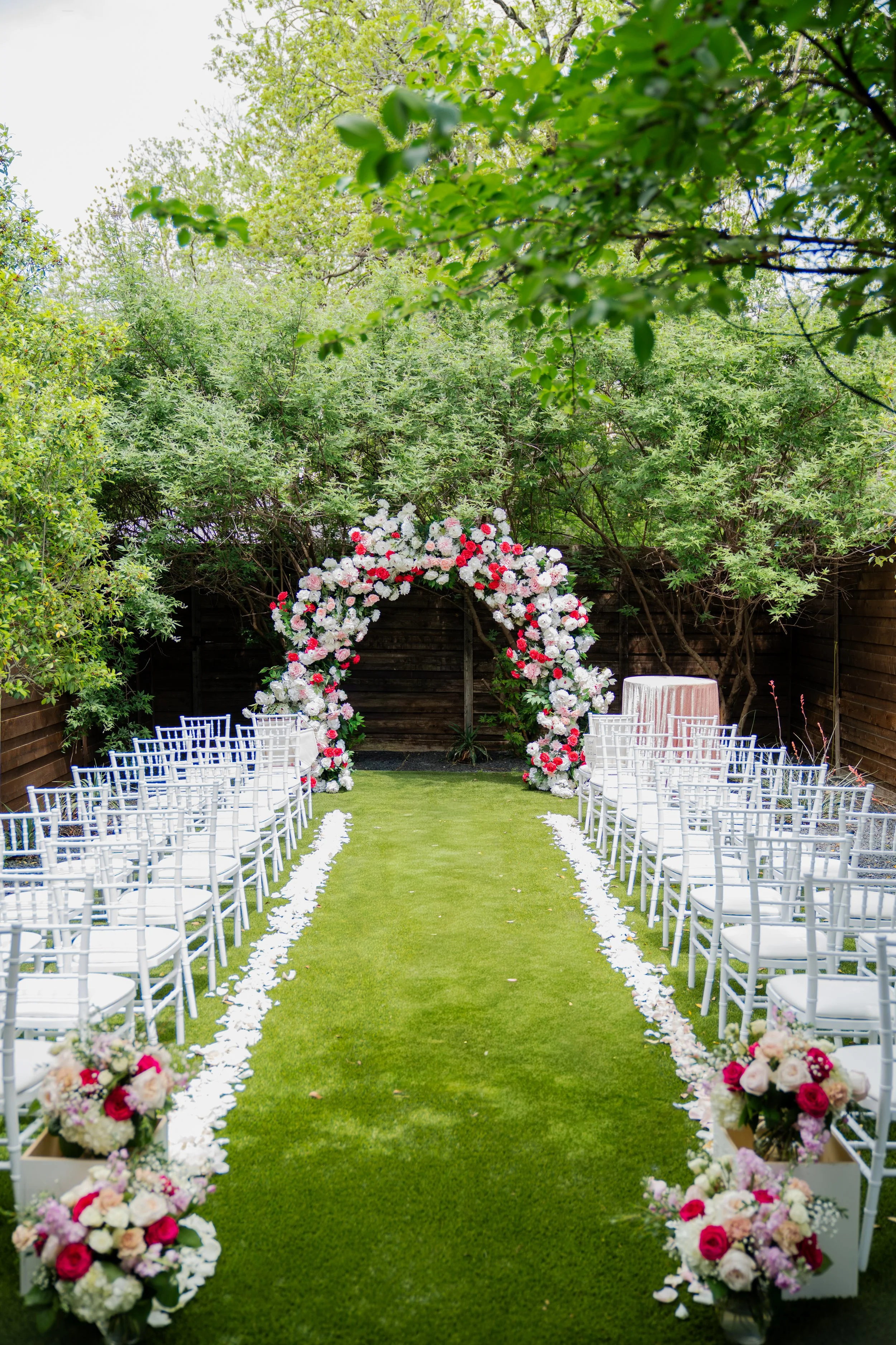 Outdoor wedding ceremony setup with white chairs arranged along a grass aisle, decorated with floral arrangements and rose petal accents, under a floral arch and surrounded by lush green trees.