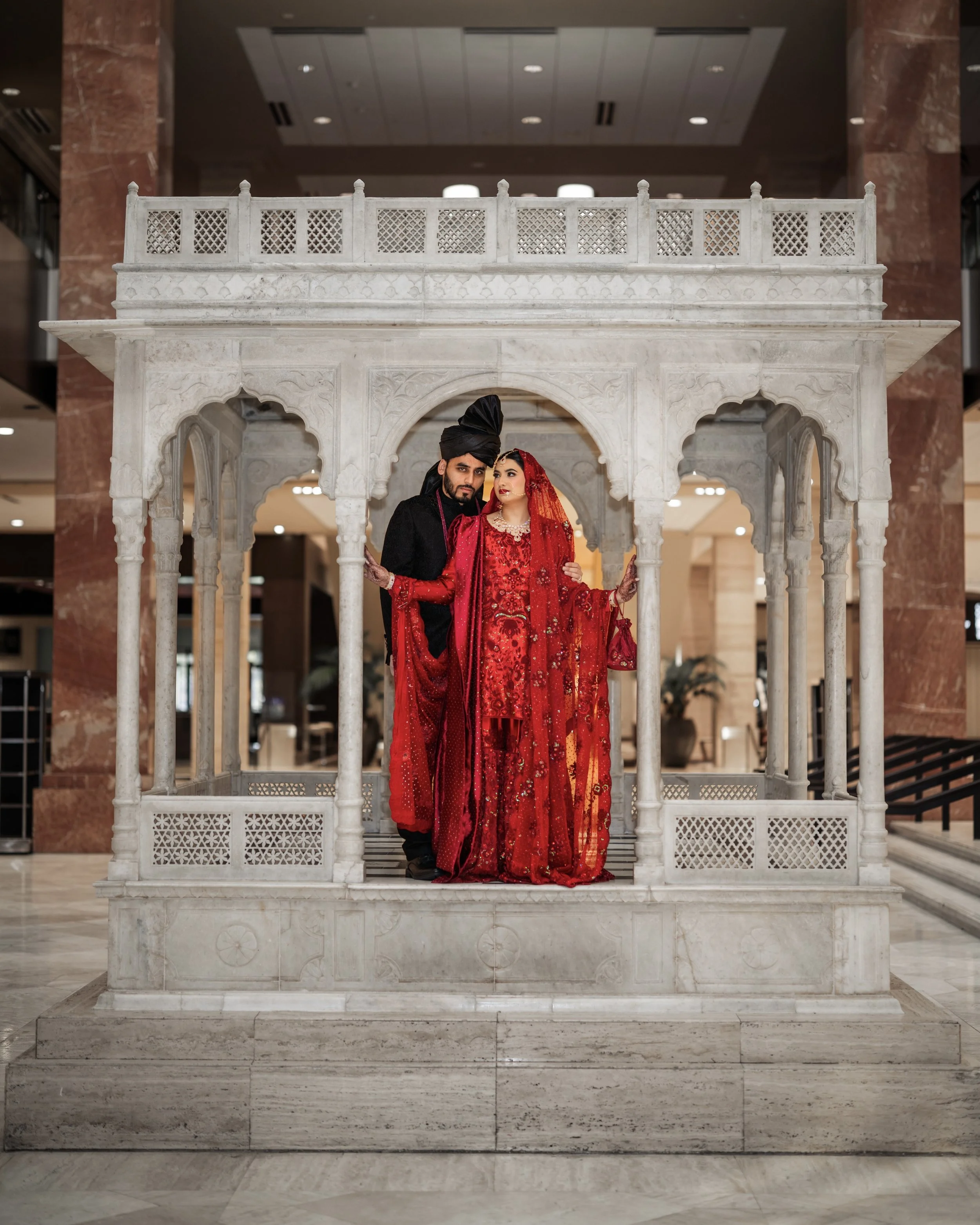 A couple dressed in traditional Indian wedding attire standing inside a decorative white marble pavilion in a hotel lobby.