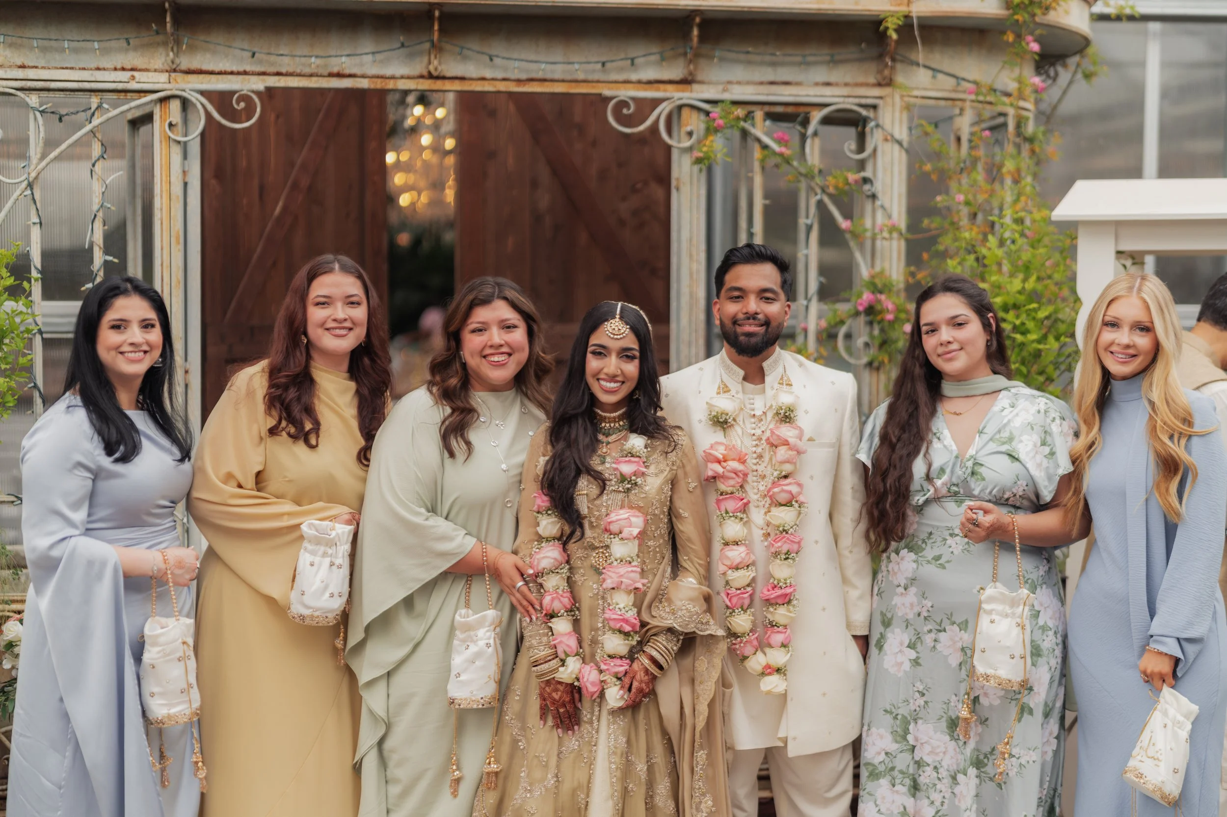 Group of seven people dressed in traditional Indian attire, standing outdoors with a wooden structure and pink flowers in the background.