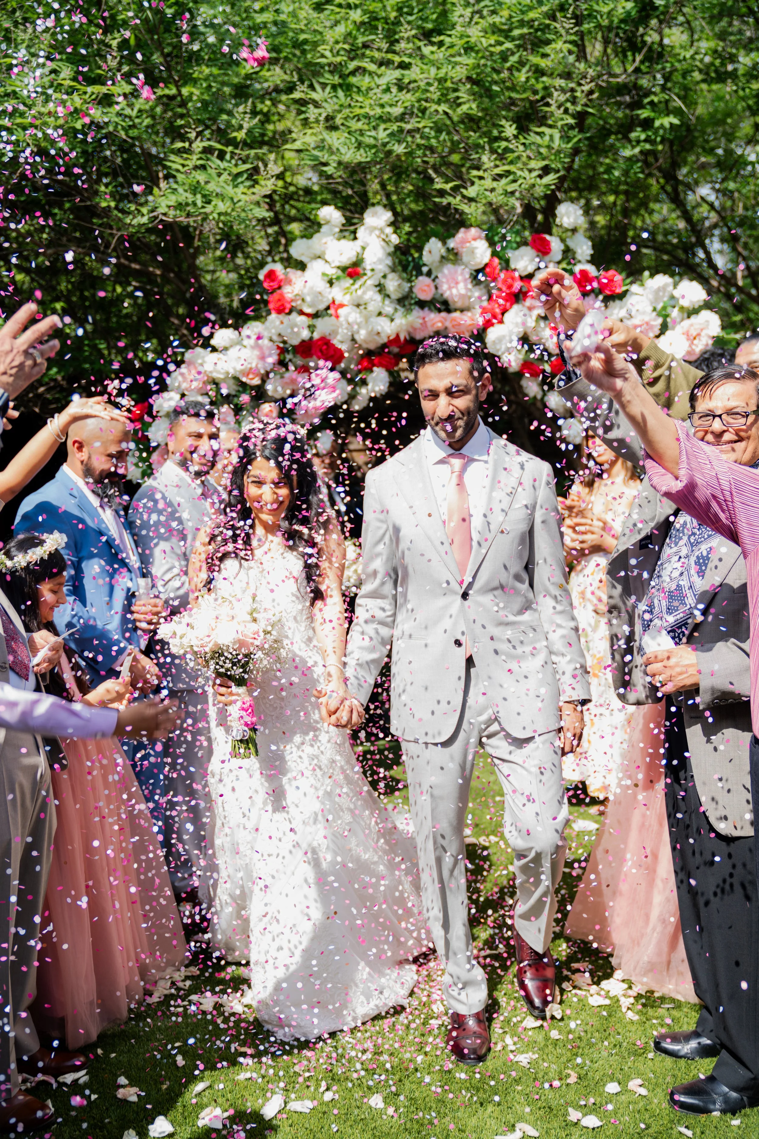 Bride and groom walking hand in hand through a shower of pink and white flower petals, surrounded by wedding guests outdoors, with a floral arch and green trees in the background.