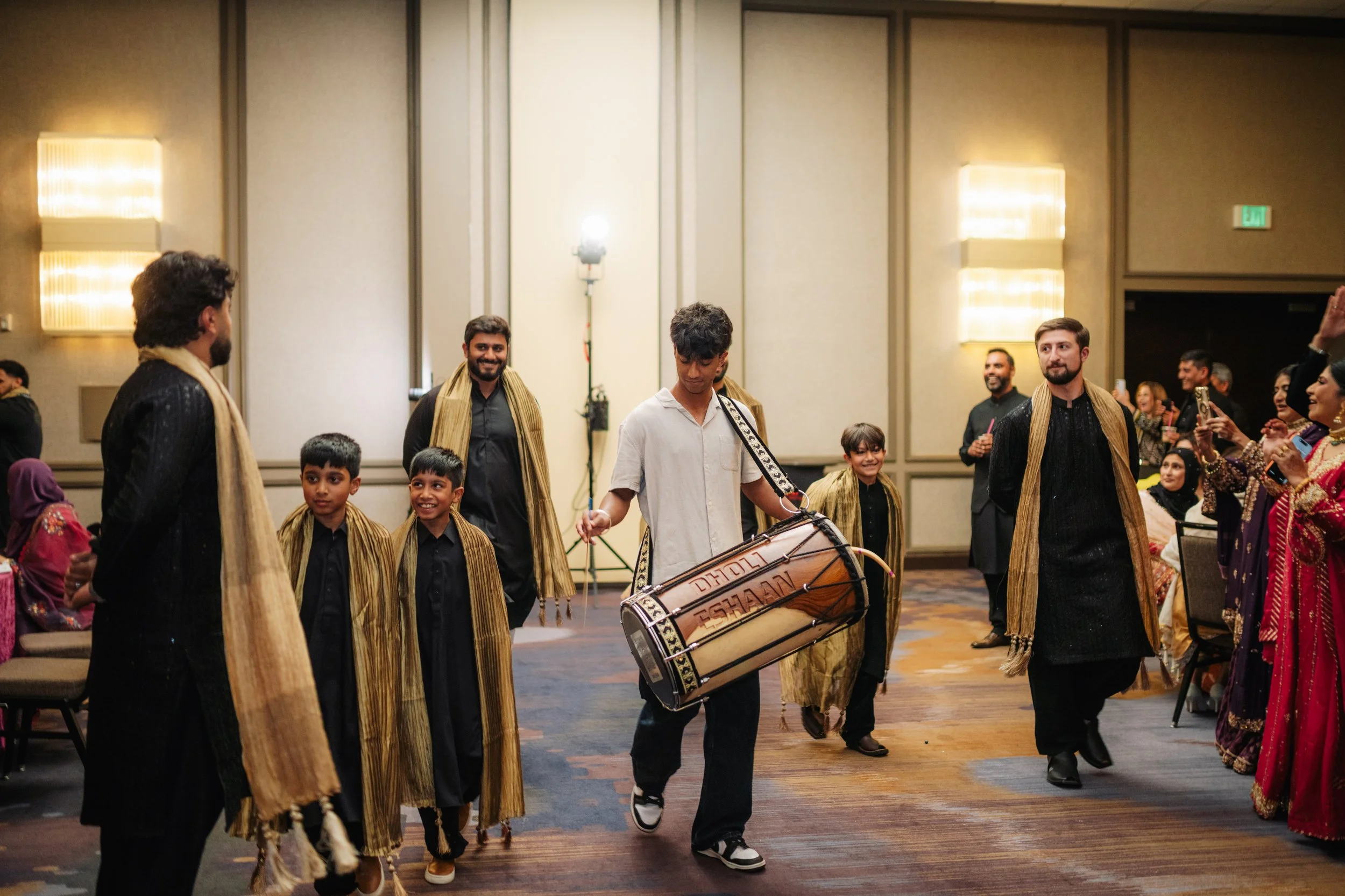 Group of people, including children and adults, dressed in traditional South Asian clothing, walking indoors at a cultural event or celebration, some playing drums and others smiling and watching.