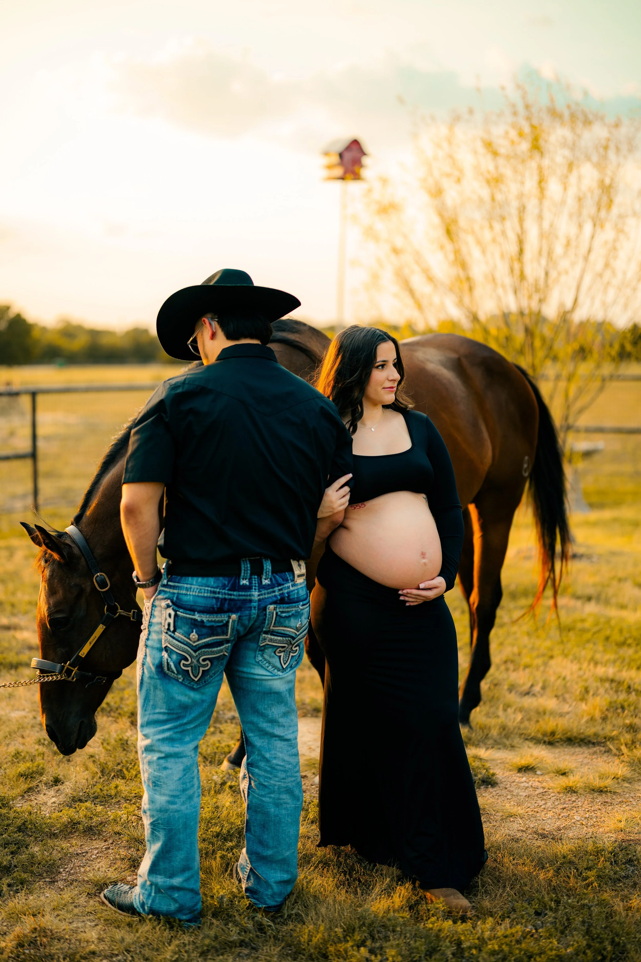A pregnant woman stands outside with a man and a horse during sunset in a rural setting with a fence and a tree in the background.