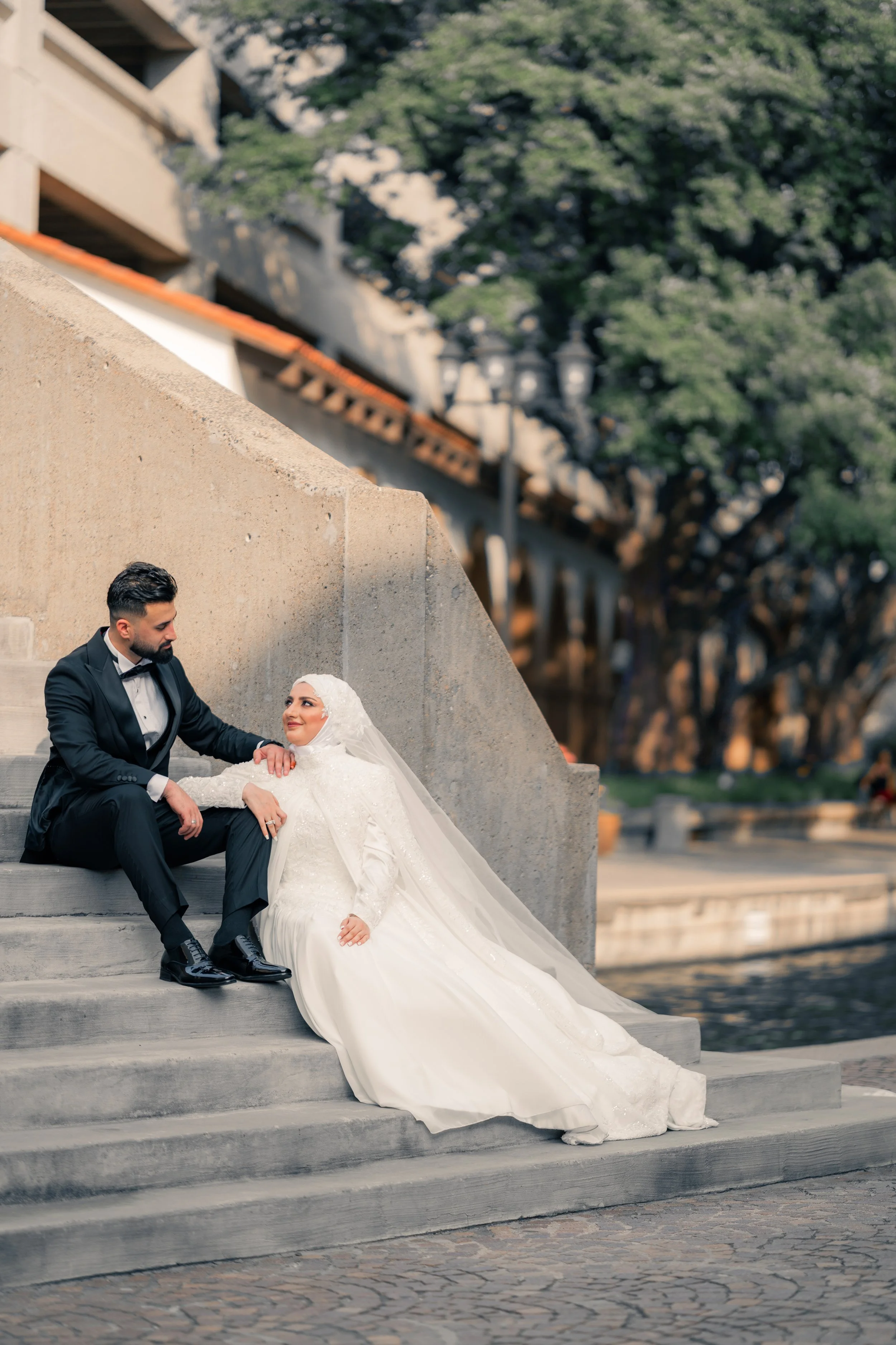 Bride and groom on steps outdoors, the bride in a white wedding dress and hijab, the groom in a black tuxedo, look at each other affectionately.
