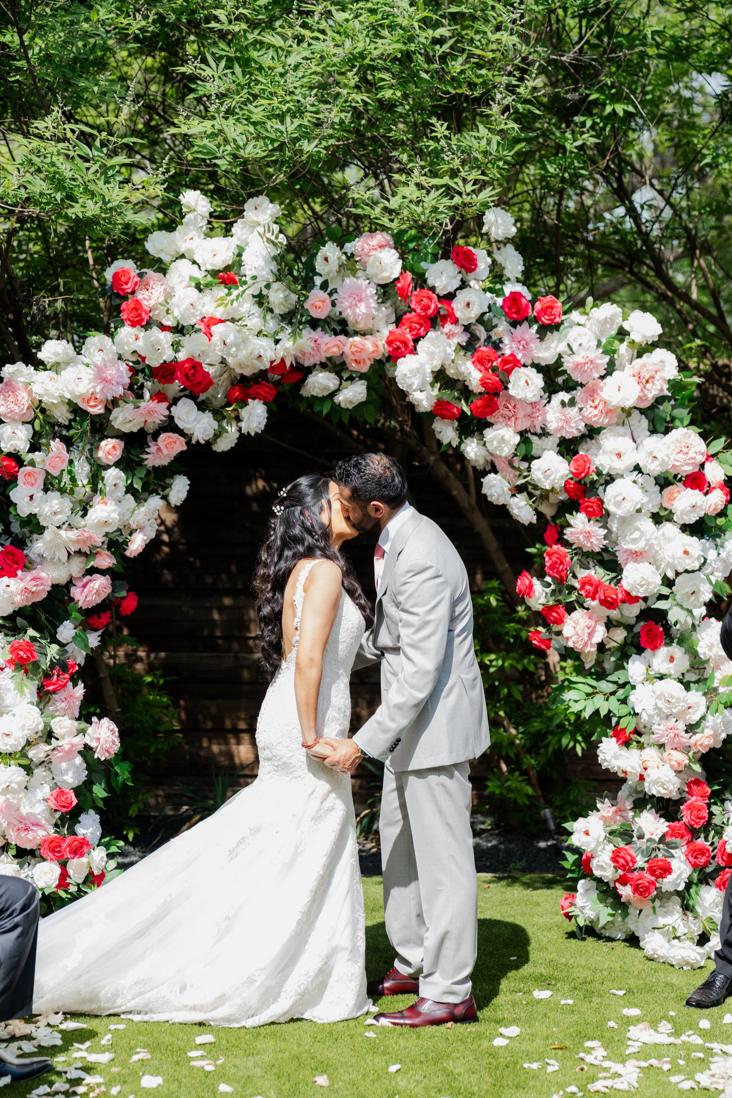 A bride and groom kissing under a floral archway at their outdoor wedding ceremony. The arch is decorated with white, pink, and red flowers, and there are rose petals scattered on the grass.