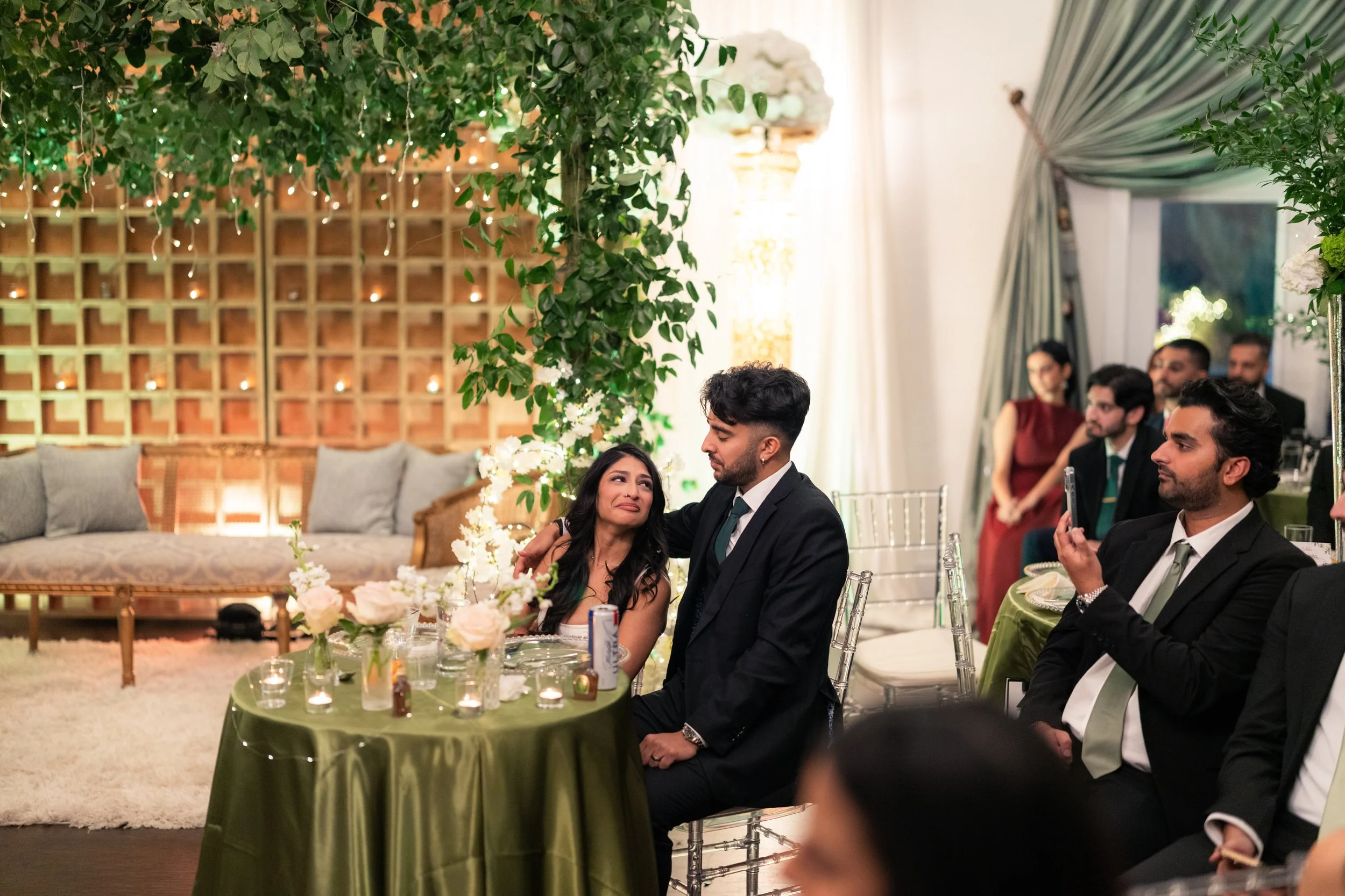 A woman tears up while sitting next to a man in a black suit at a decorated wedding reception, with guests seated nearby and floral arrangements on the table.
