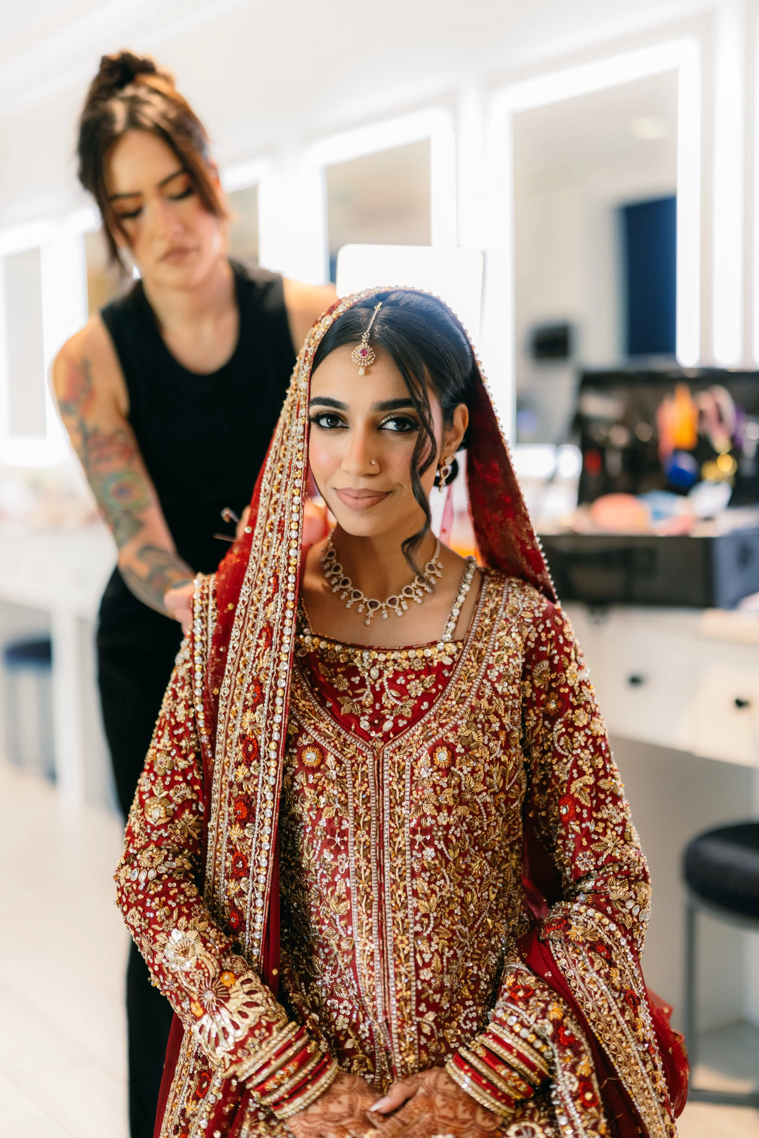 A woman dressed in traditional Indian bridal attire with intricate red and gold embroidery, jewelry, and henna on her hands, being prepared by a stylist in a bright room.