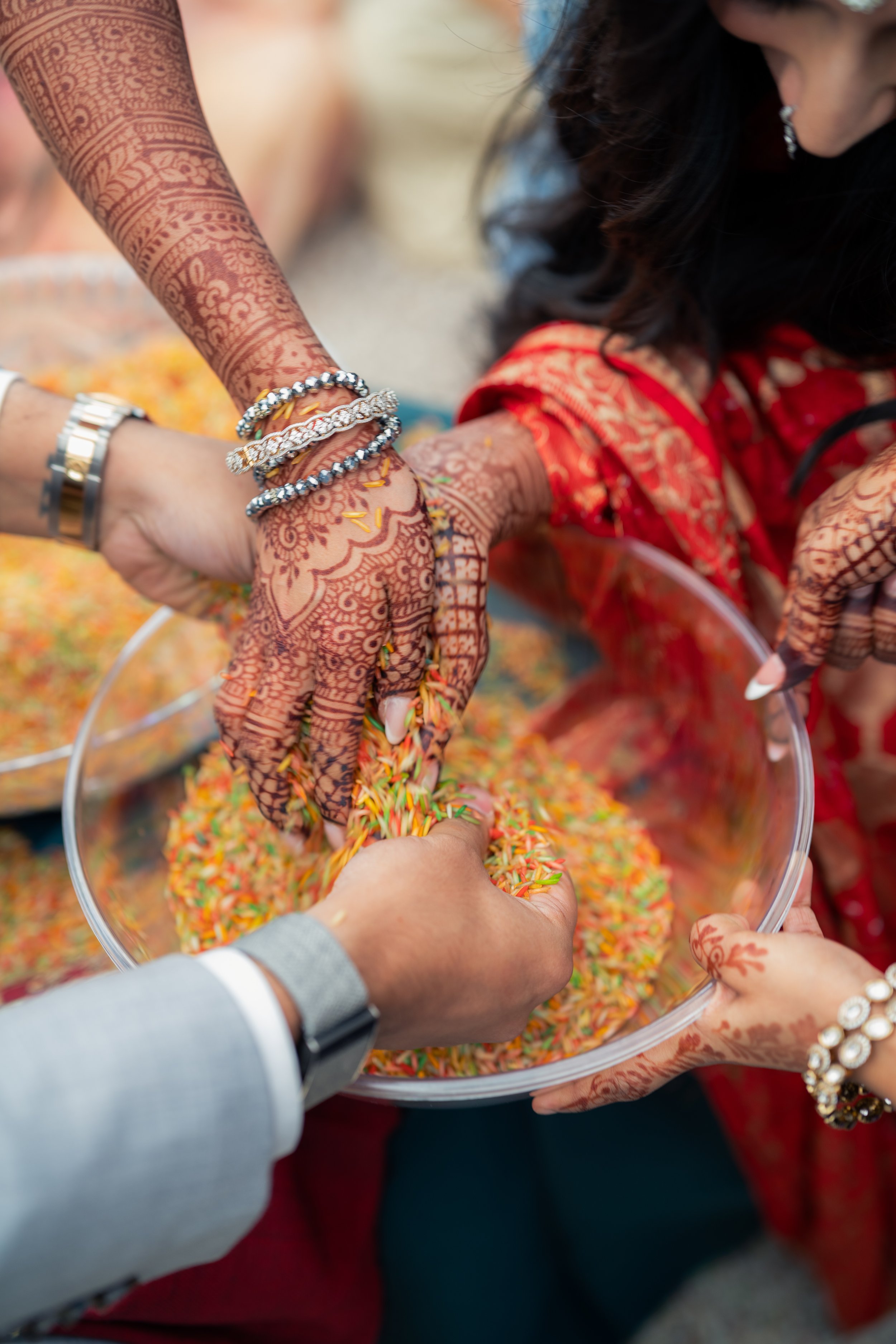 Multiple hands with henna designs are reaching into a large glass bowl filled with colorful rice, participating in a traditional cultural or celebratory activity.