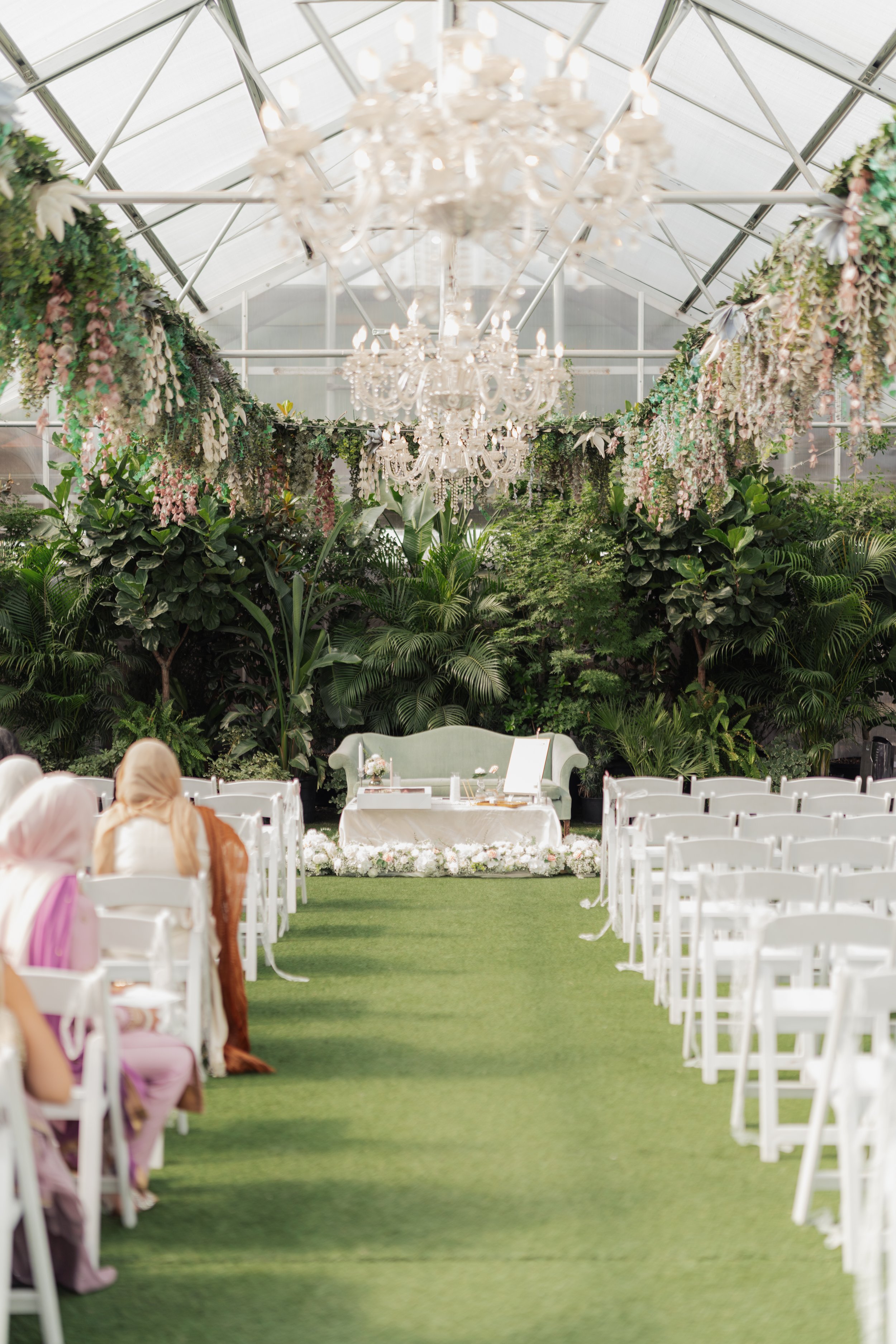 Indoor wedding ceremony setup inside a glass-enclosed venue with white chairs, a white sofa, and floral decorations, featuring hanging chandeliers and greenery.