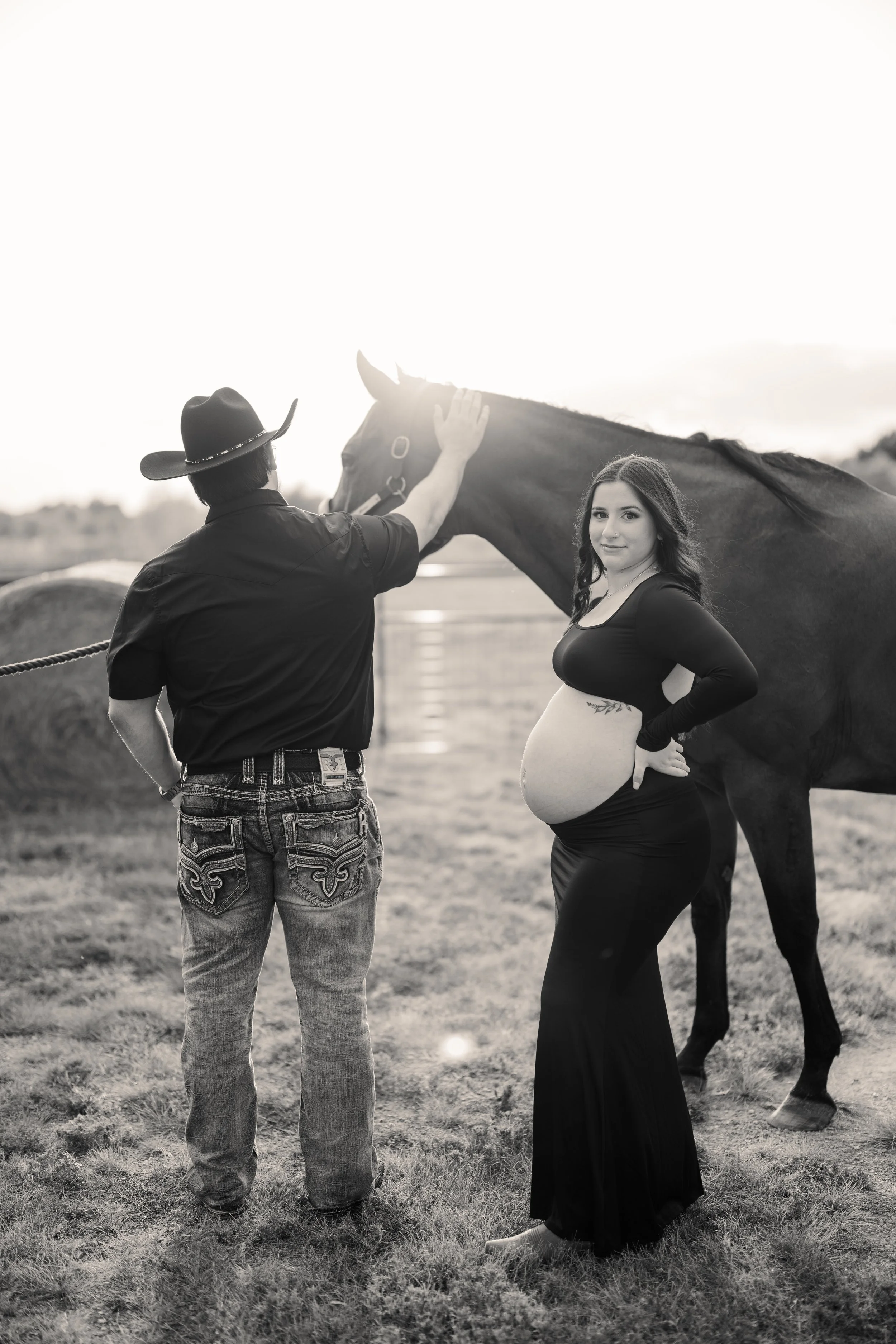 A pregnant woman in a long black dress standing with a cowgirl hat and a man in a cowboy shirt and jeans, outdoors on grass, with a horse nearby, black and white photo.