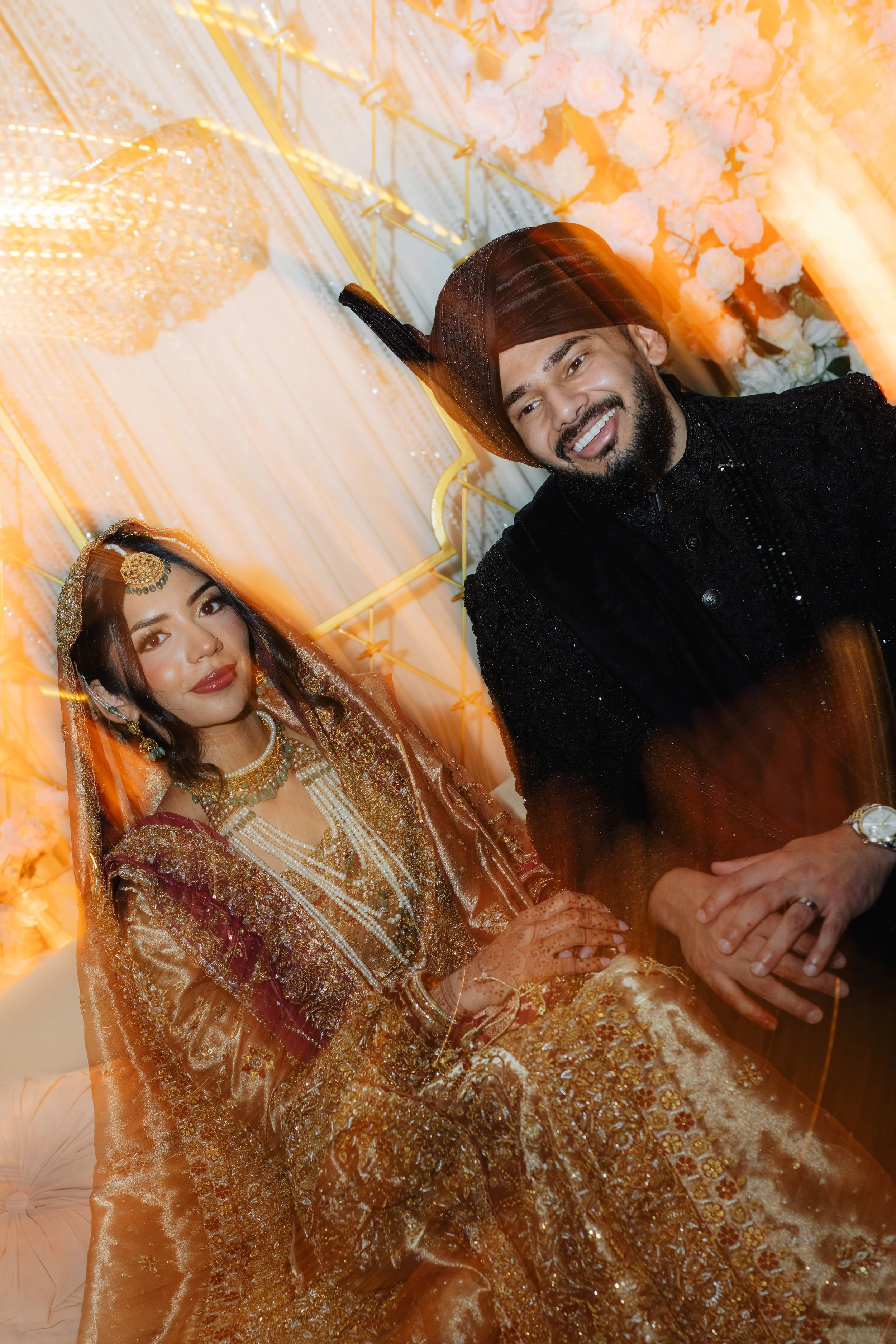 A smiling man and woman in traditional, ornate Indian wedding attire, seated in front of floral and decorative backdrop.