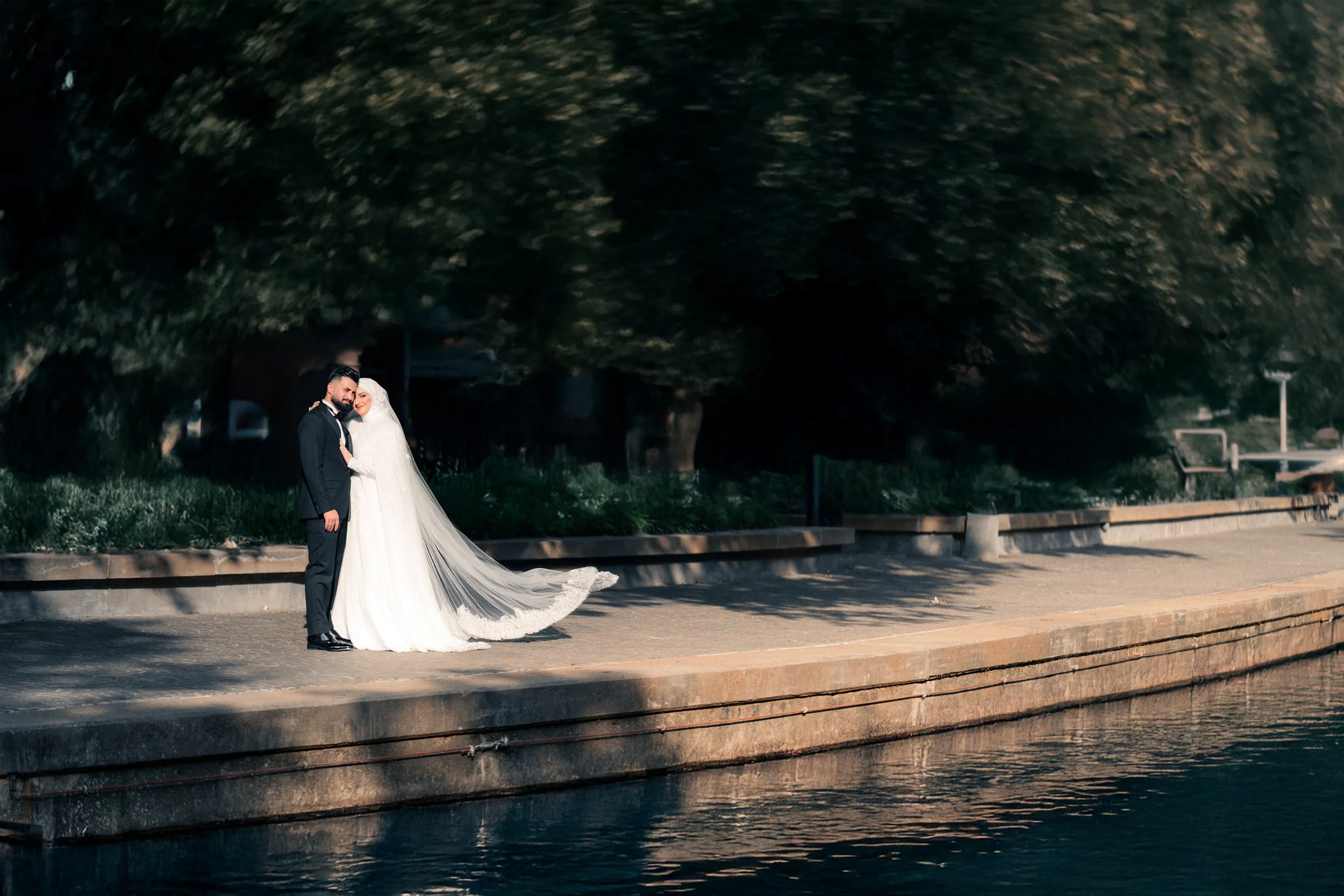 A bride and groom standing together on a waterfront promenade, with the bride wearing a white wedding gown and veil, and the groom in a black tuxedo, under large trees.