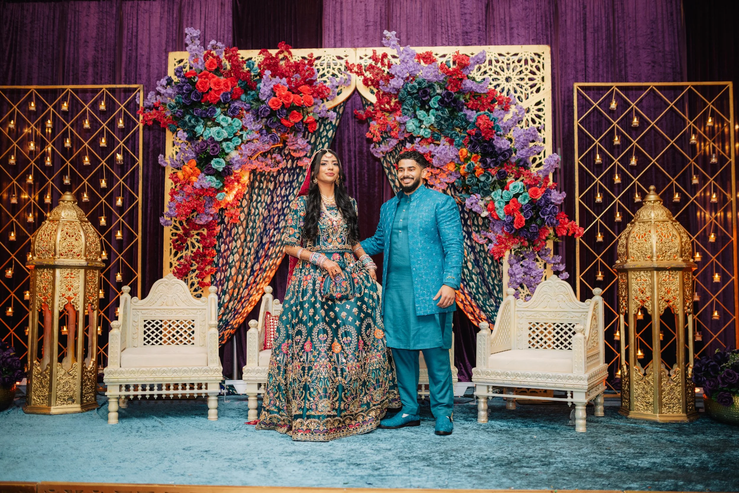 A couple in traditional Indian wedding attire standing on a decorated stage with floral arrangements and candles.