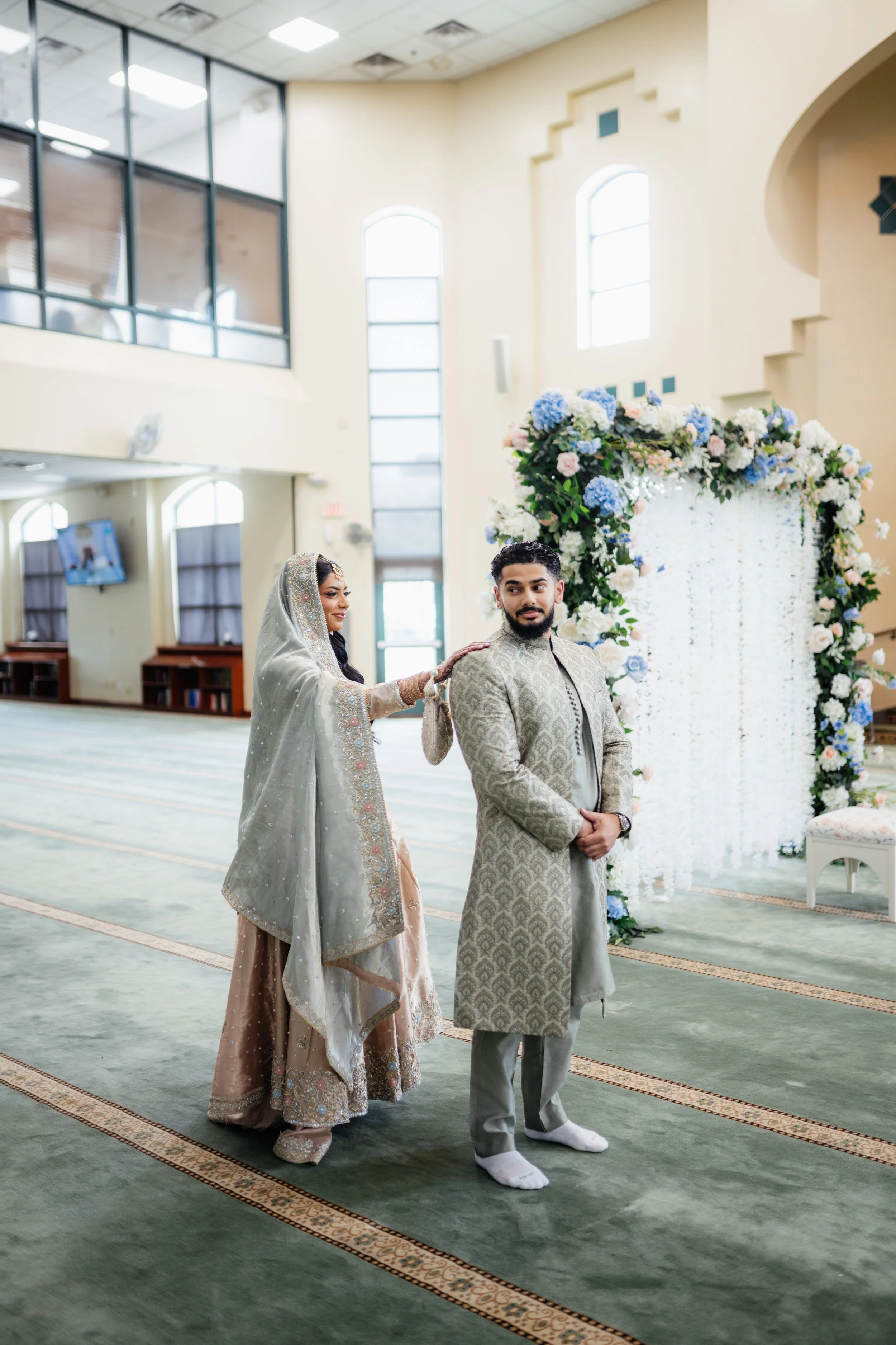 A bride and groom standing inside a church decorated with a flower arch of white, pink, and blue flowers. The bride is wearing a traditional South Asian bridal outfit with intricate embroidery, and the groom is dressed in a matching traditional outfi