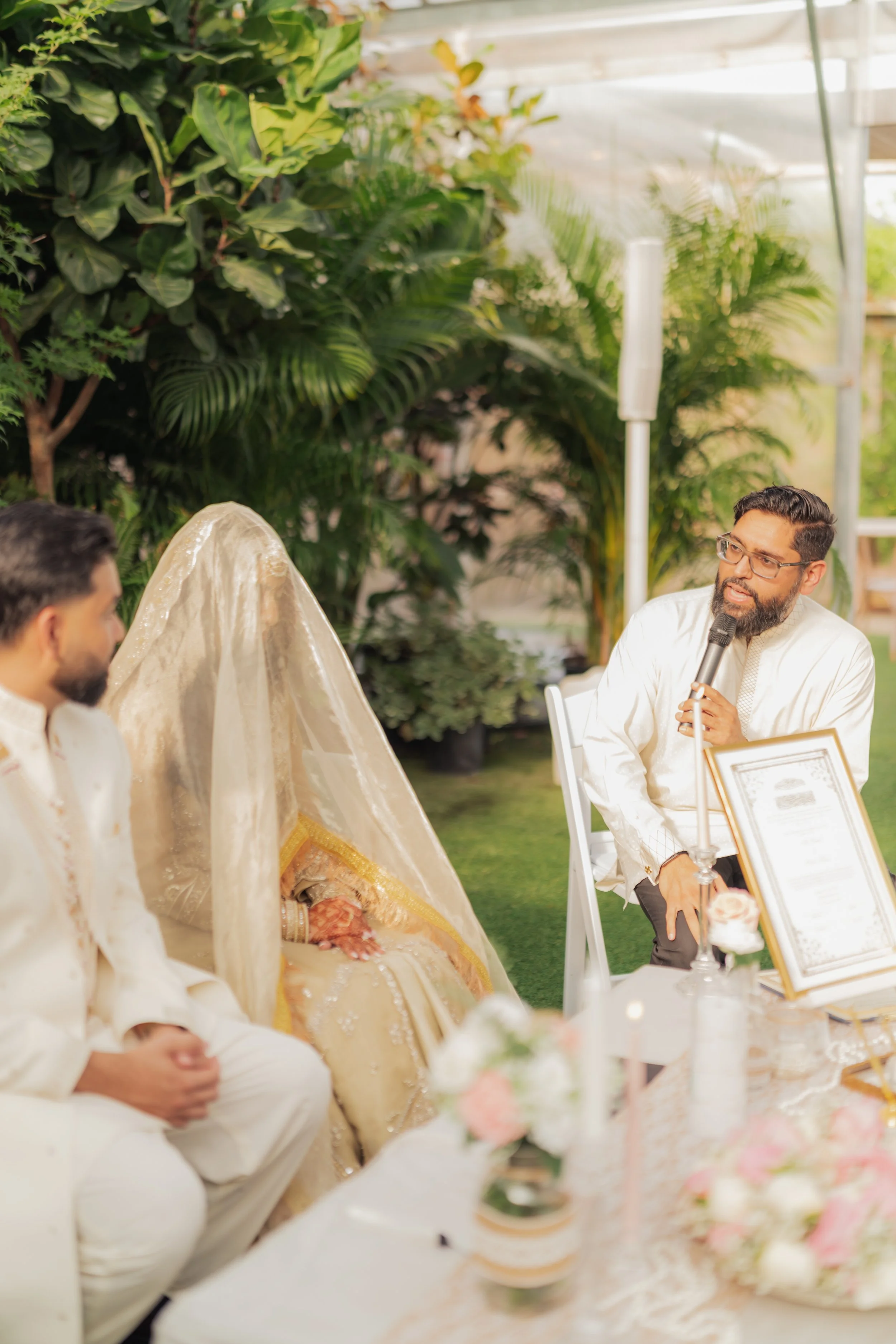 A group of people attending a wedding celebration outdoors, with a lush green background, and a man speaking into a microphone.