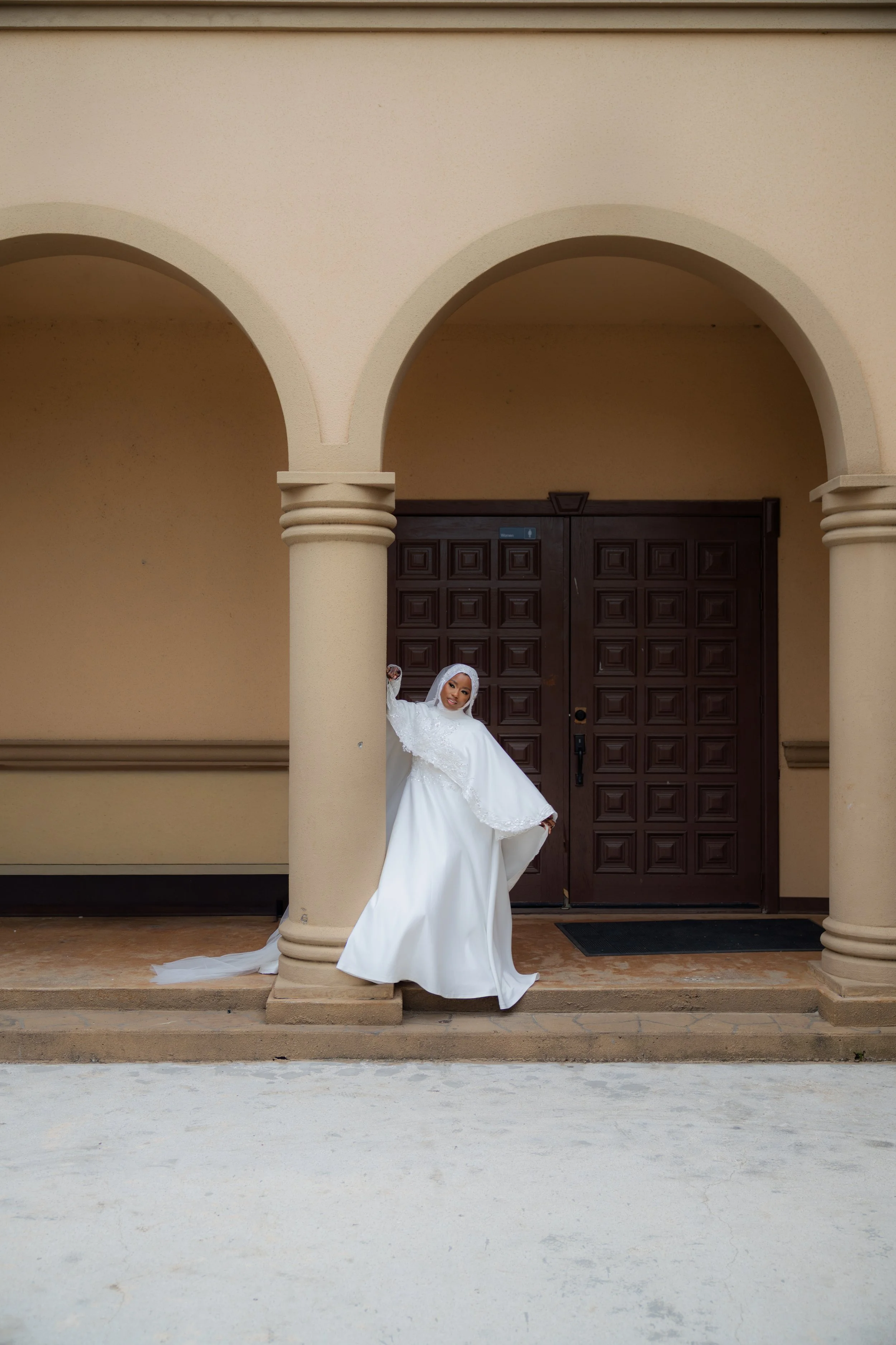 Brunette woman in white wedding dress standing between two beige columns in front of a wooden door.