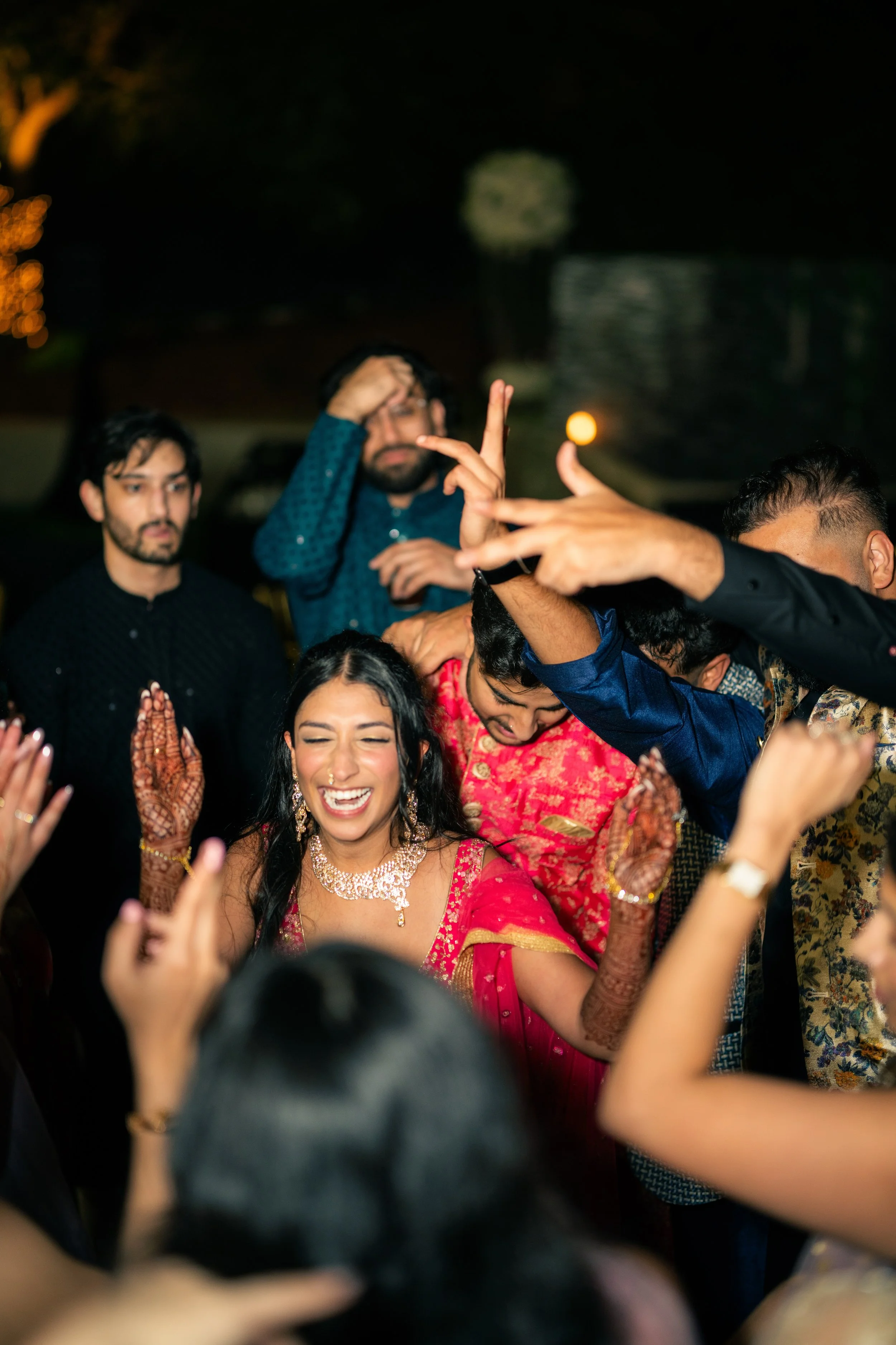 A woman in traditional South Asian attire and jewelry, smiling and enjoying herself at a celebration with a group of people, some of whom are wearing colorful clothes, outdoors at night.