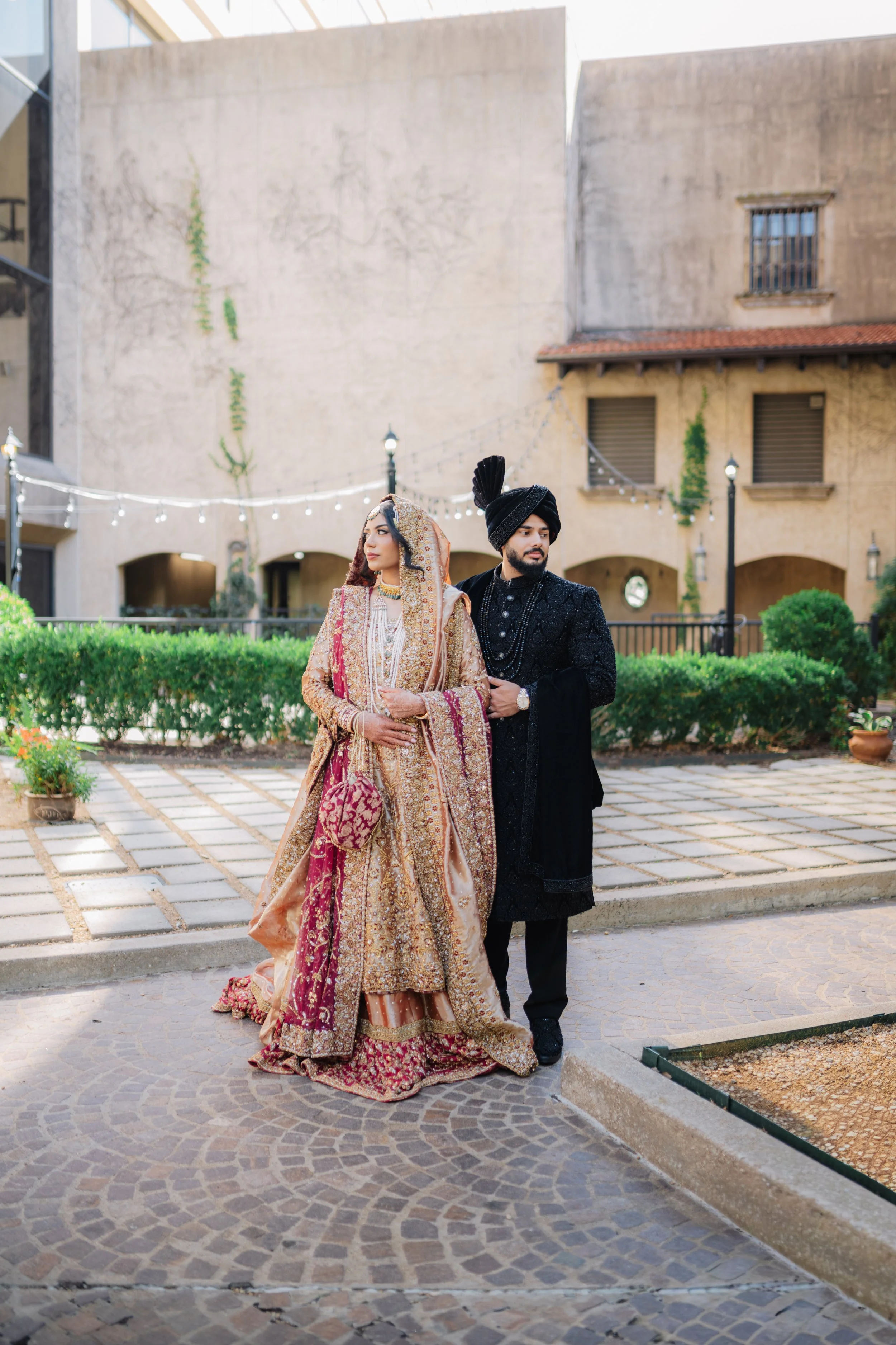A South Asian bride and groom standing outside on a paved area with a shrub and a beige building in the background, dressed in traditional wedding attire.