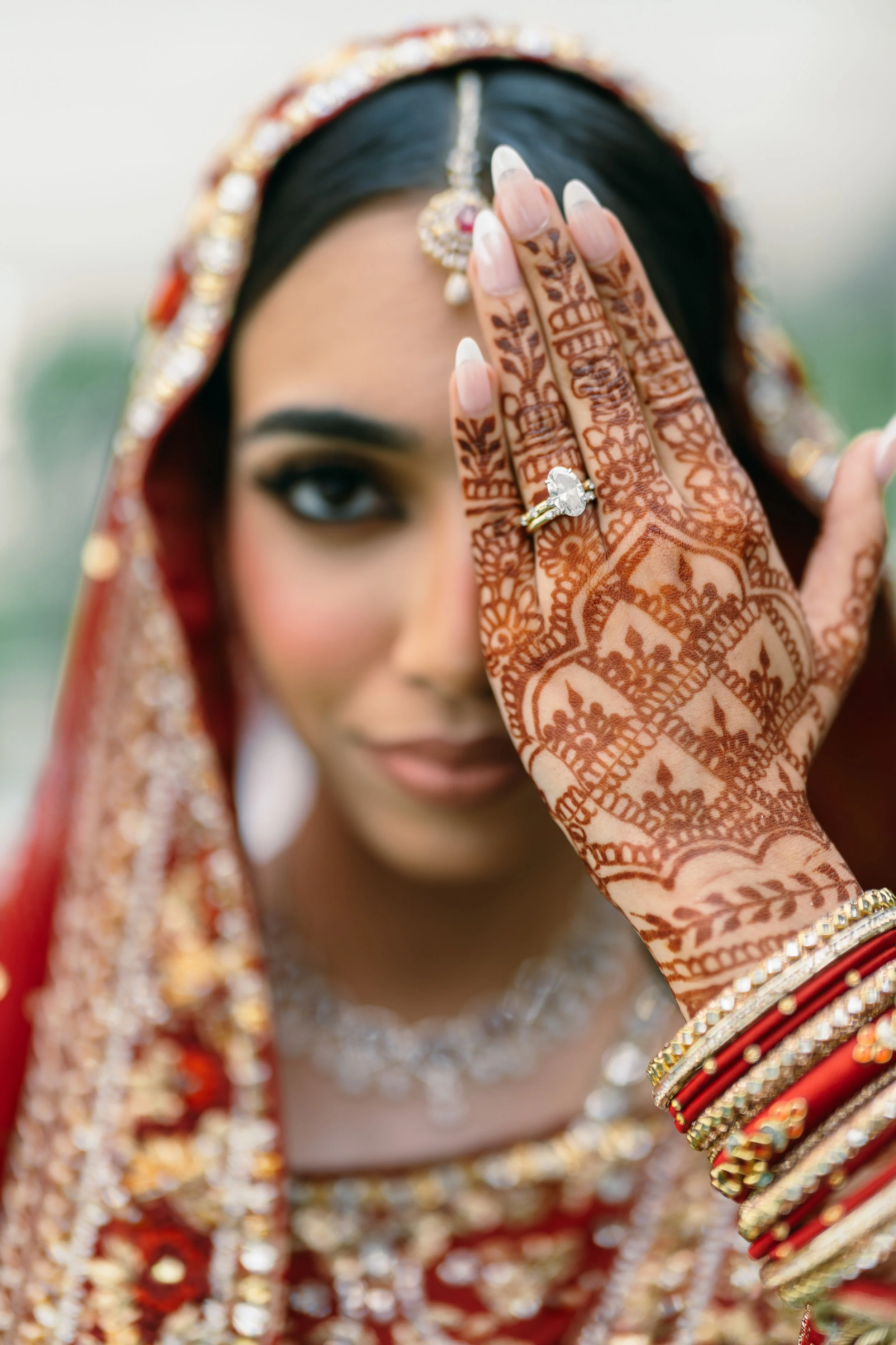 A woman in traditional Indian wedding attire with intricate henna designs on her hand, an engagement ring, and decorated jewelry, covering part of her face with her hand.