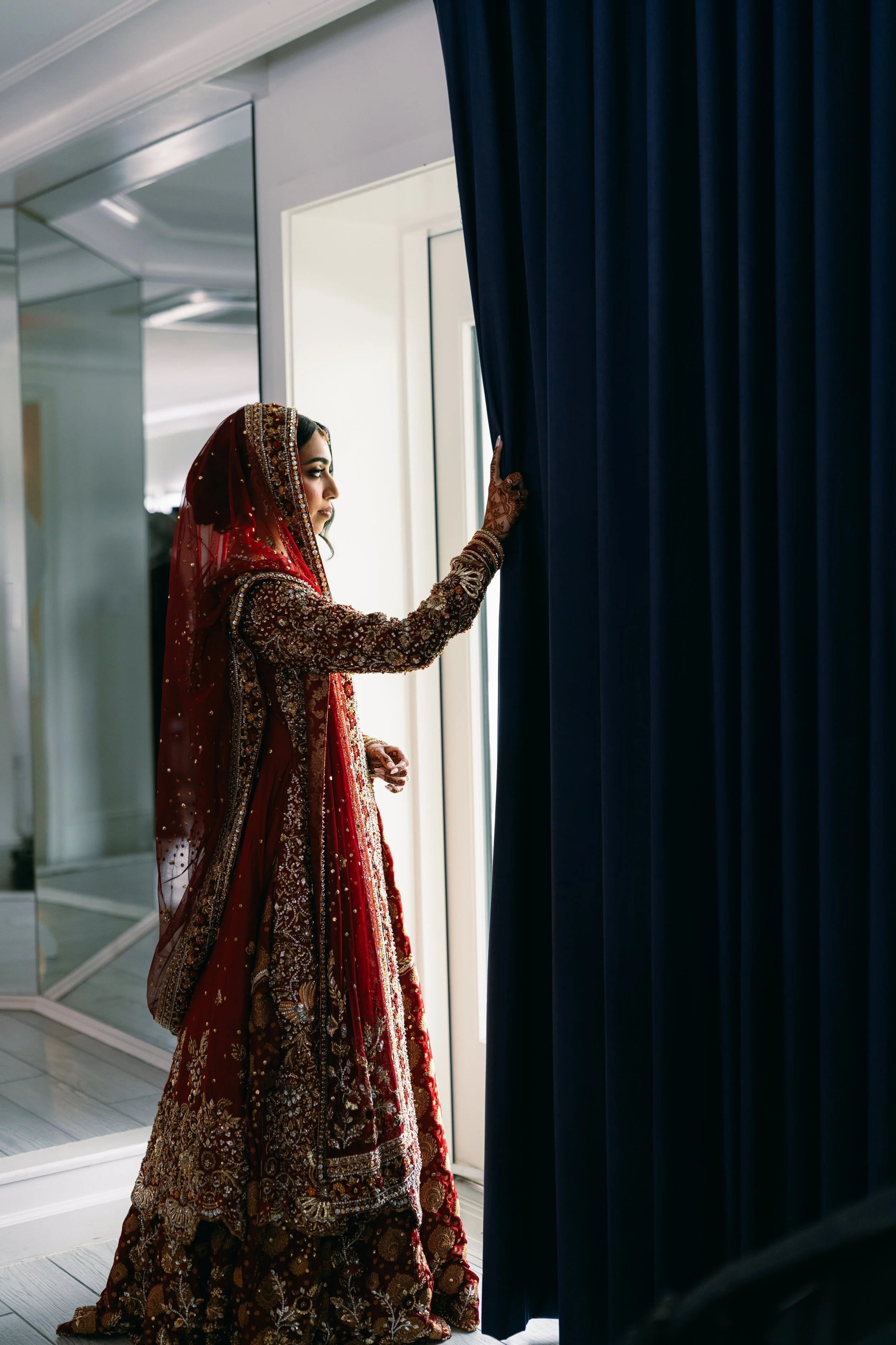 A woman dressed in a traditional red and gold embroidered dress with a matching veil is gazing out a window while gently touching the curtain.