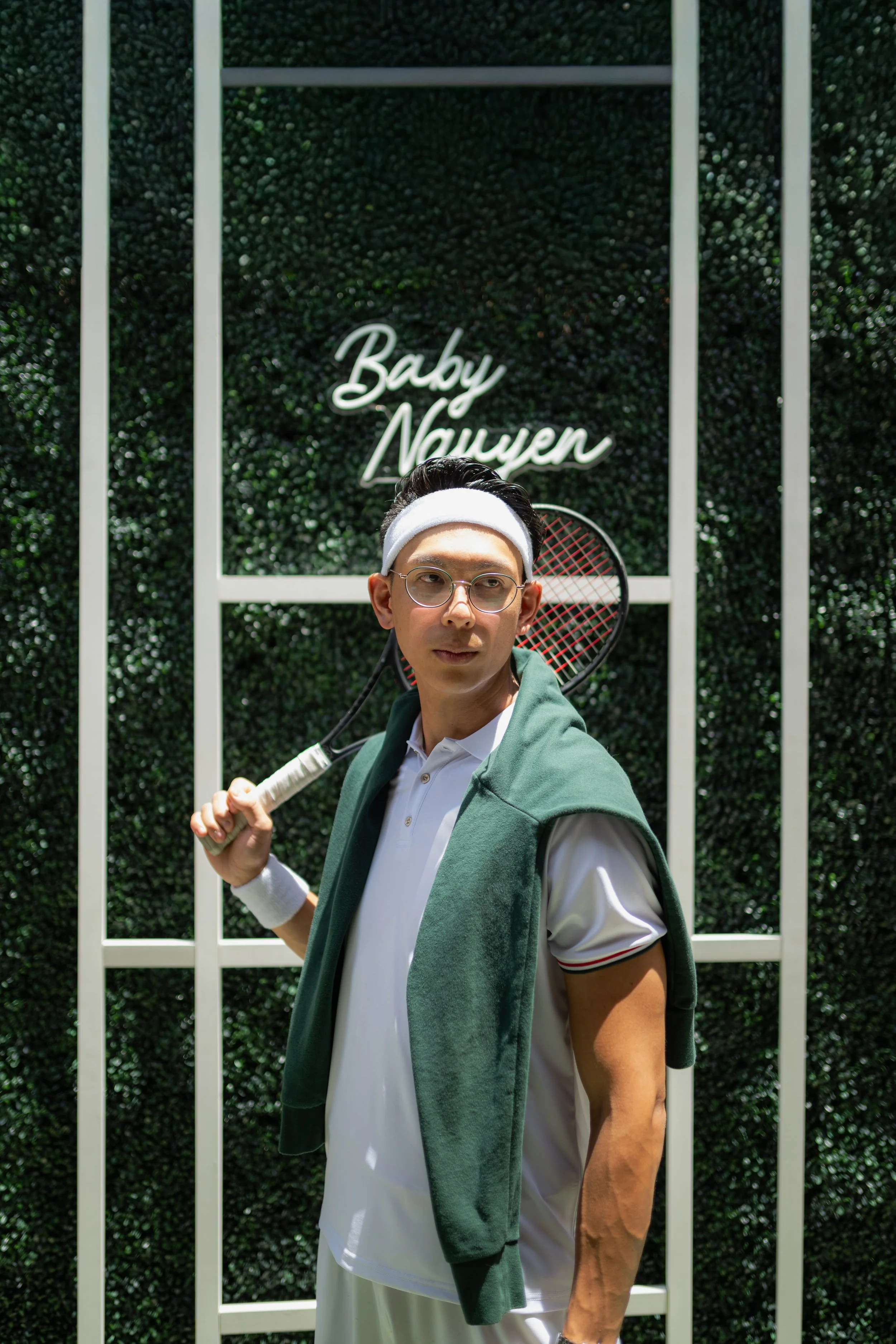 A man standing outdoors in front of a green hedge wall with a neon sign that reads 'Baby Nouwen'; he is holding a tennis racket over his shoulder, wearing glasses, a white headband, a white polo shirt, and a green jacket draped over his shoulders.