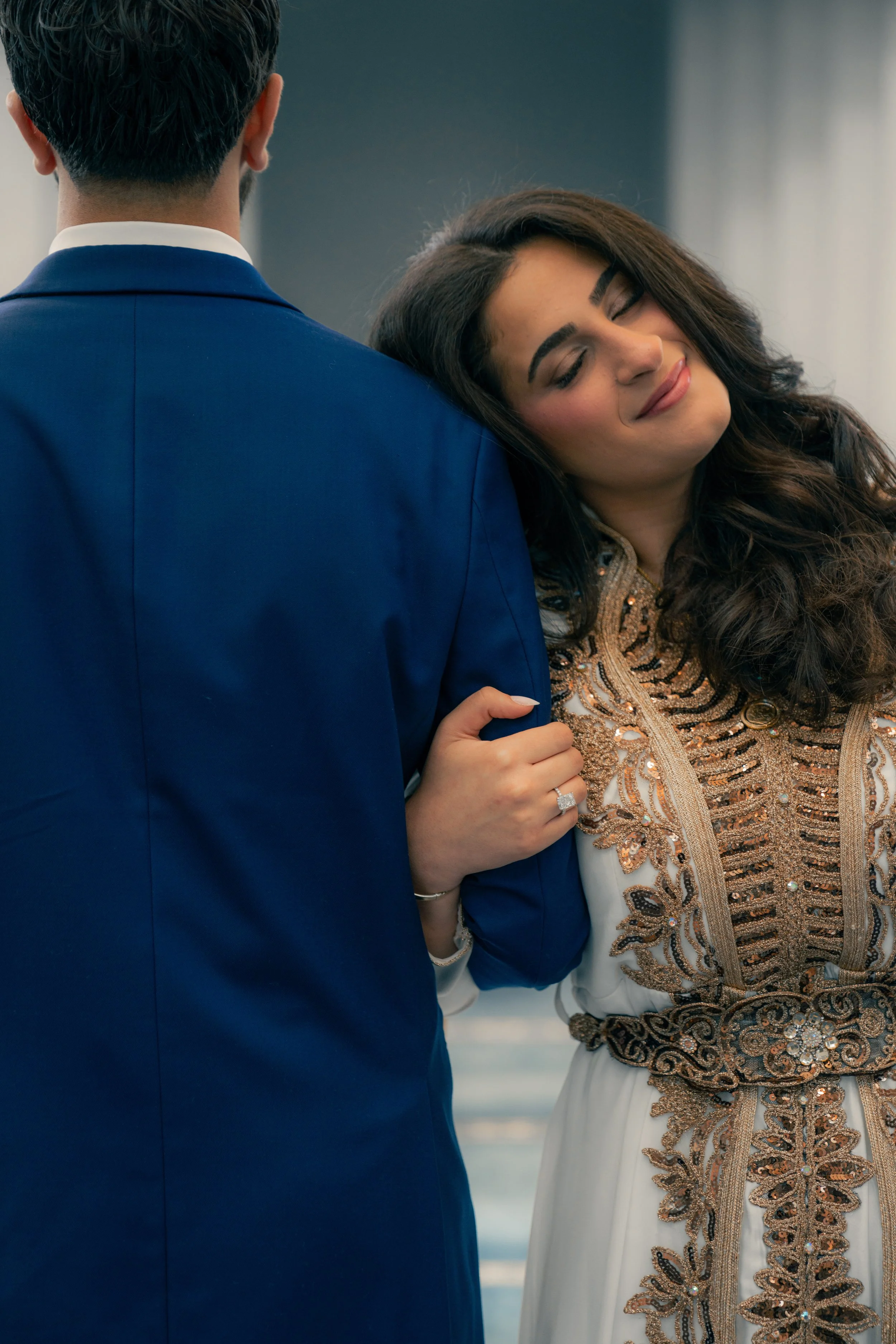A woman in an elegant dress with intricate gold and black embroidery leans her head on a man's shoulder, who is wearing a blue suit. The woman has long dark hair and a ring on her finger.