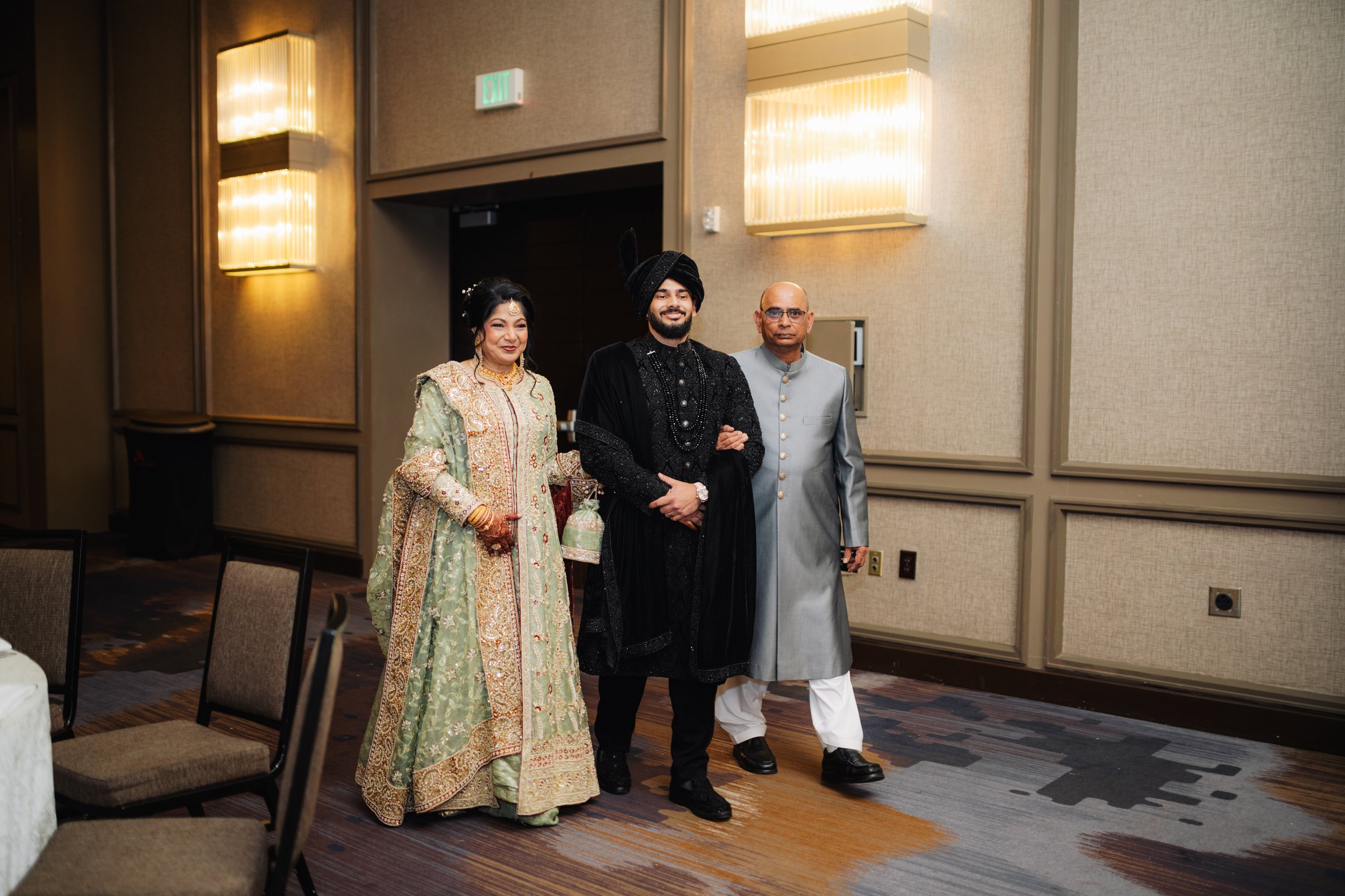 Three people dressed in ornate traditional Indian attire standing together in a banquet hall, smiling. The woman is wearing a green and gold embroidered dress, the man in the middle is in a black sherwani with a turban, and the man on the right is in