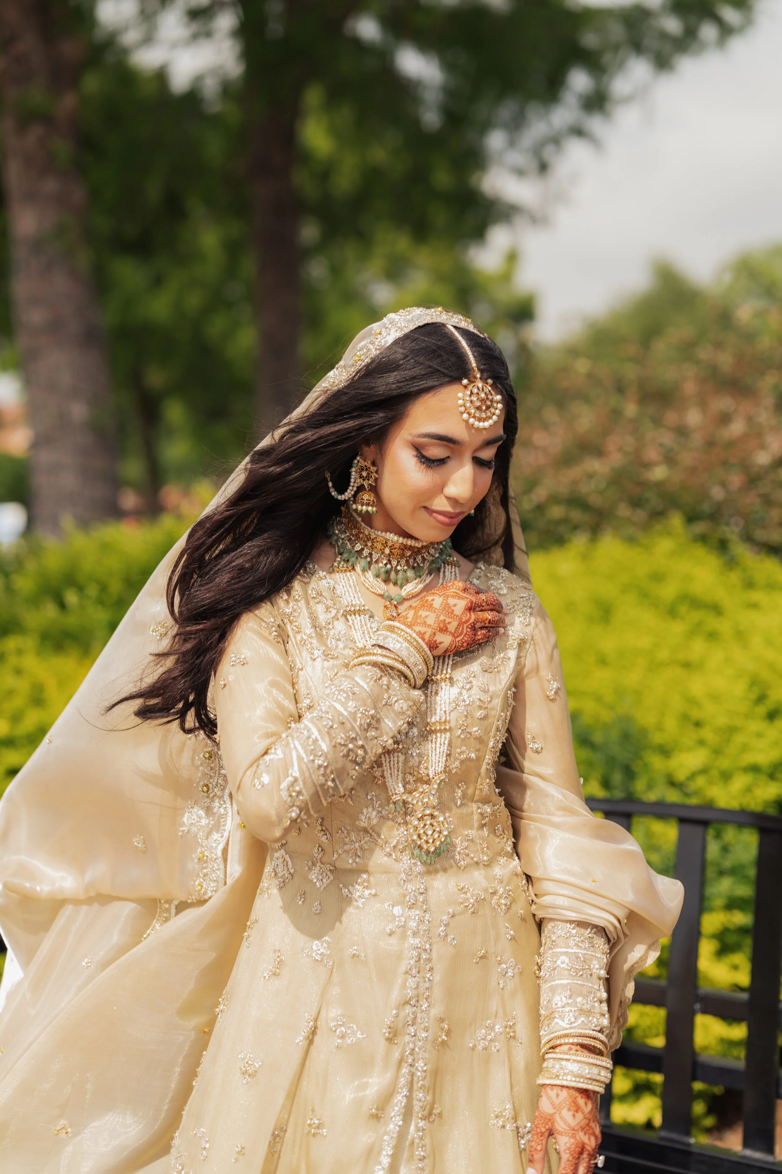 A woman dressed in traditional South Asian bridal attire, wearing an ornate cream-colored dress with embroidery, jewelry including earrings, necklace, and headpiece, outdoors with greenery and trees in the background.