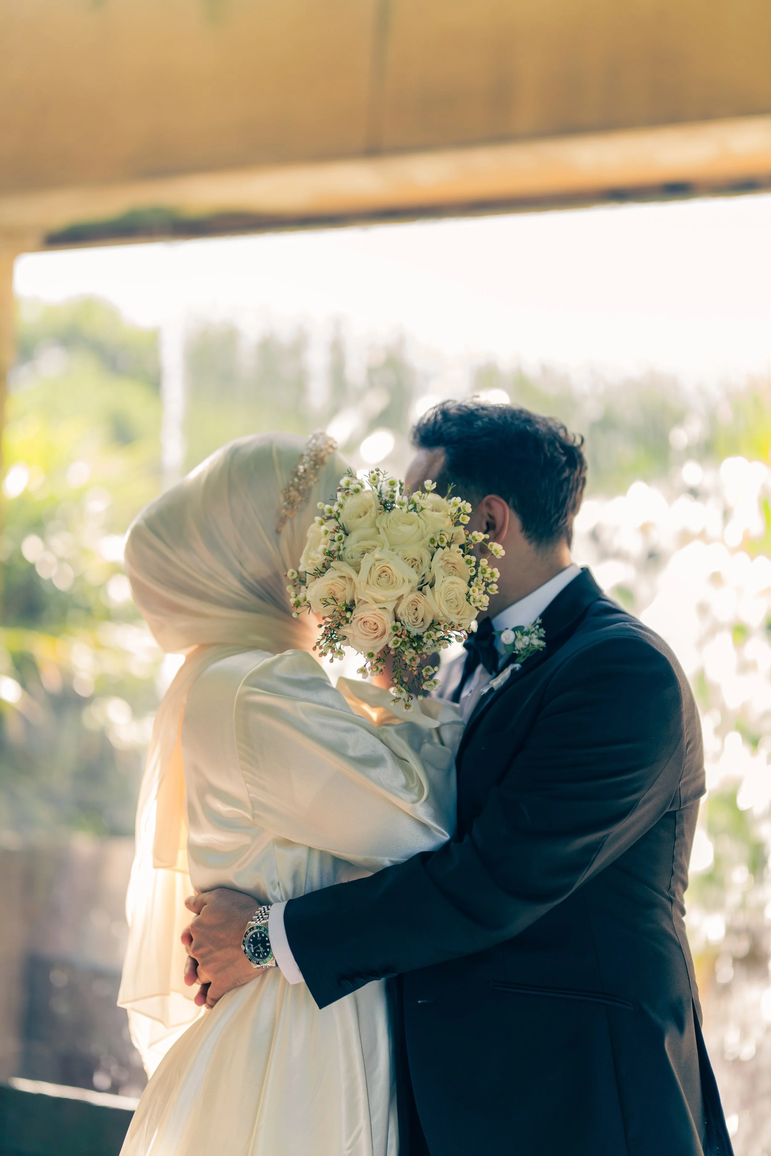 A bride and groom embracing during their wedding, with the bride holding a bouquet of white roses, in front of a bright outdoor background.