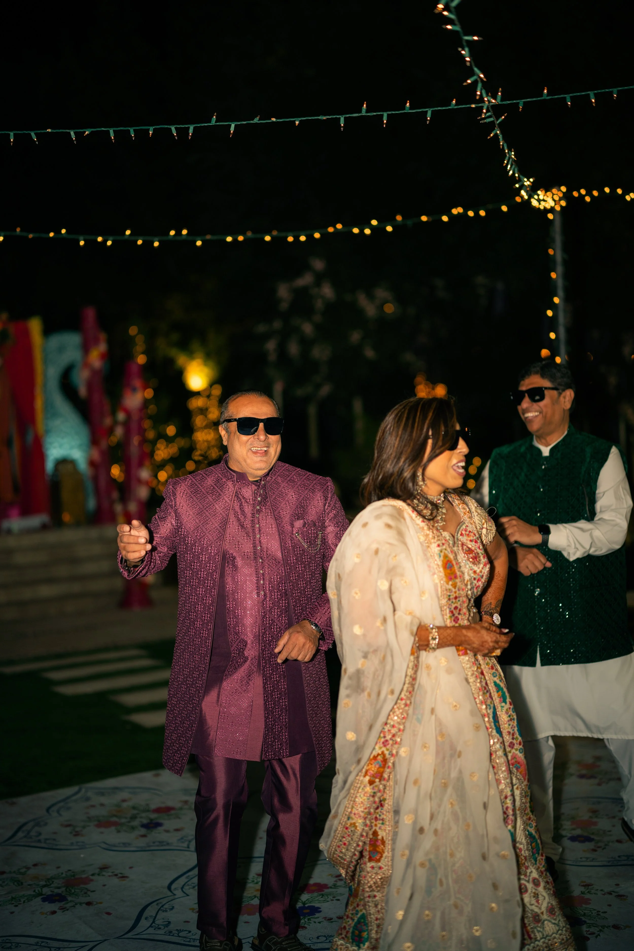 Three people dressed in traditional Indian attire are celebrating at night under string lights, with a decorative background.
